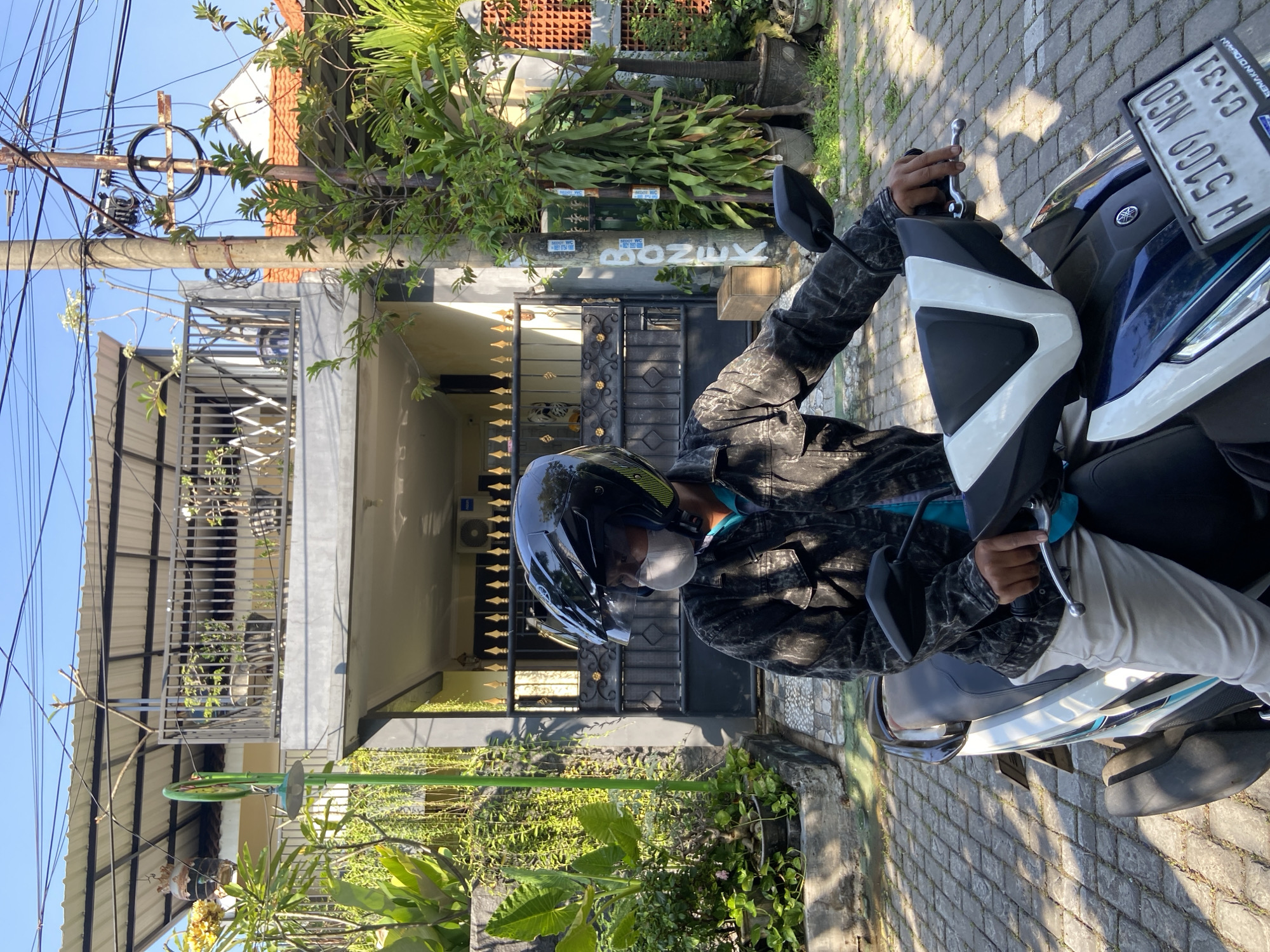 A person wearing a black helmet, a light-colored mask, and a dark patterned jacket is seated on a white and blue motorcycle. The person is looking to the left. The motorcycle is parked on a brick-paved surface. In the background, there is a two-story building with a gray facade and a white roof. A black metal gate with decorative gold accents is in front of the building. Lush green plants are visible on both sides of the entrance and on a balcony on the second floor. A utility pole with numerous wires is also present to the right of the building. The license plate of the motorcycle reads "W 5109 NGO C1-31". The scene appears to be set in a residential area during daylight, with clear blue skies. The specific location is in the city of Gelam, Indonesia.