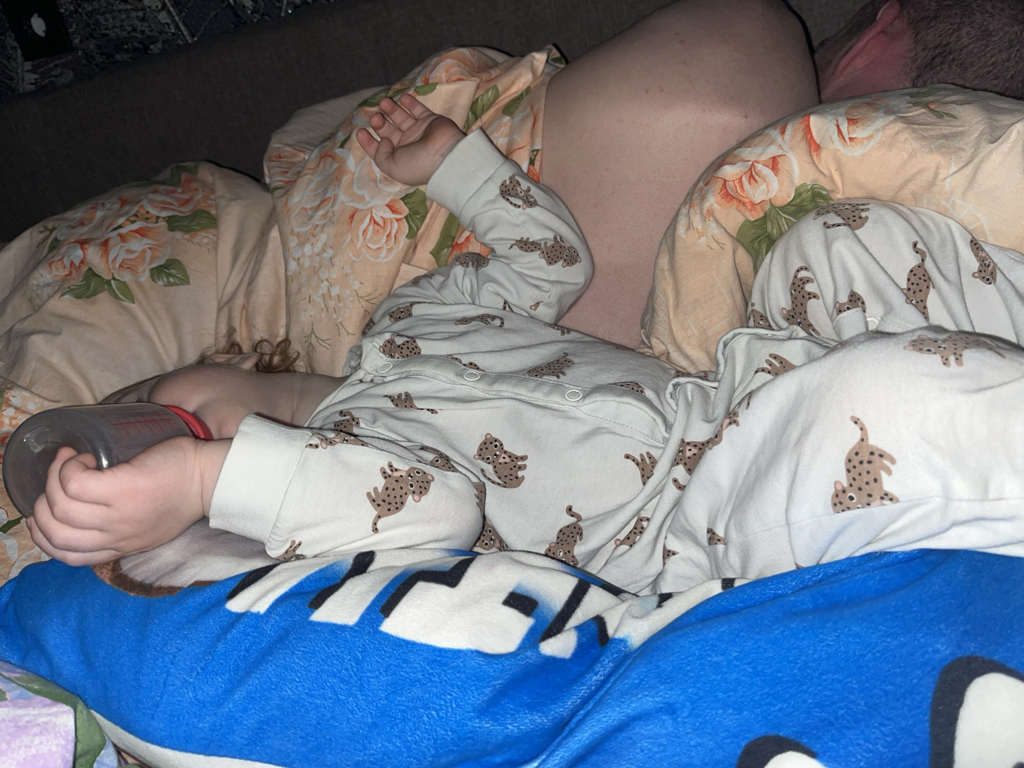 A baby wearing a white onesie with a brown leopard print is lying on a bed with peach and floral print bedding. The baby is holding a bottle with a red cap in their left hand. A blue and white blanket with a cartoon design is partially covering the baby. An adult's shoulder and ear are visible behind the baby's head. The location is unspecified, but it may or may not be Chaikovsky, Russia.
