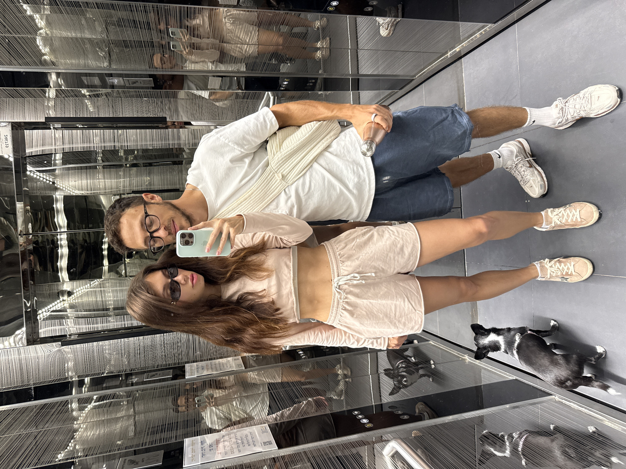 A man and a woman are in an elevator with a dog, taking a selfie. The elevator is decorated with reflective surfaces and strings. The woman wears a two-piece, light-colored outfit and sunglasses. The man wears a white t-shirt, blue shorts, and glasses. He carries a water bottle. The dog is black and white. Based on the document in Hebrew writing, the location is Tel-Aviv, Israel.