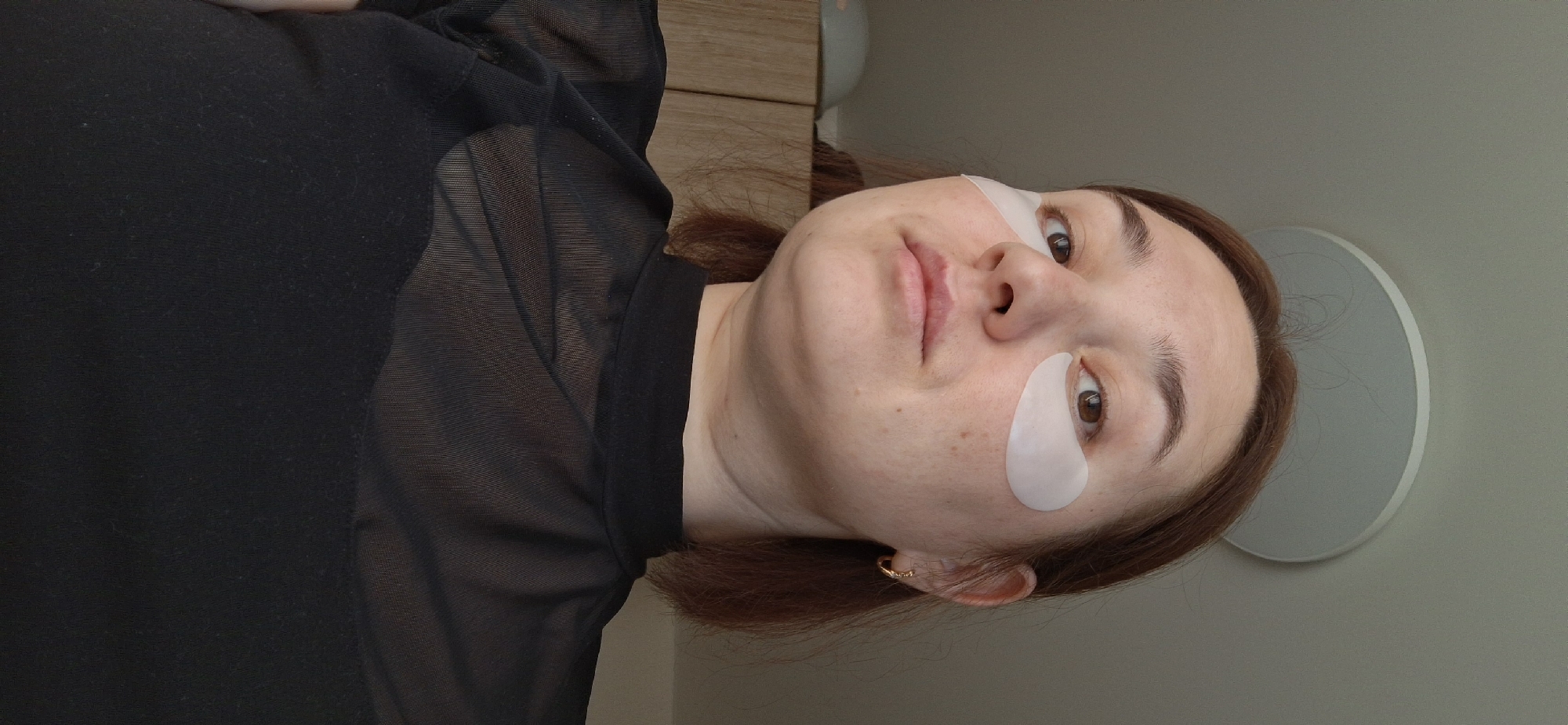 A medium shot depicts a woman lying down, facing the camera at an angle, with white under-eye patches applied. She has light skin, brown hair, and is wearing a black sheer top. The background is a neutral gray wall with a circular object partially visible, possibly a light fixture or mirror. A wooden cabinet is slightly visible to the left of her head. The location is consistent with an interior space. There is no verifiable information to determine this photo was taken in the city of Chaikovsky, Russia.