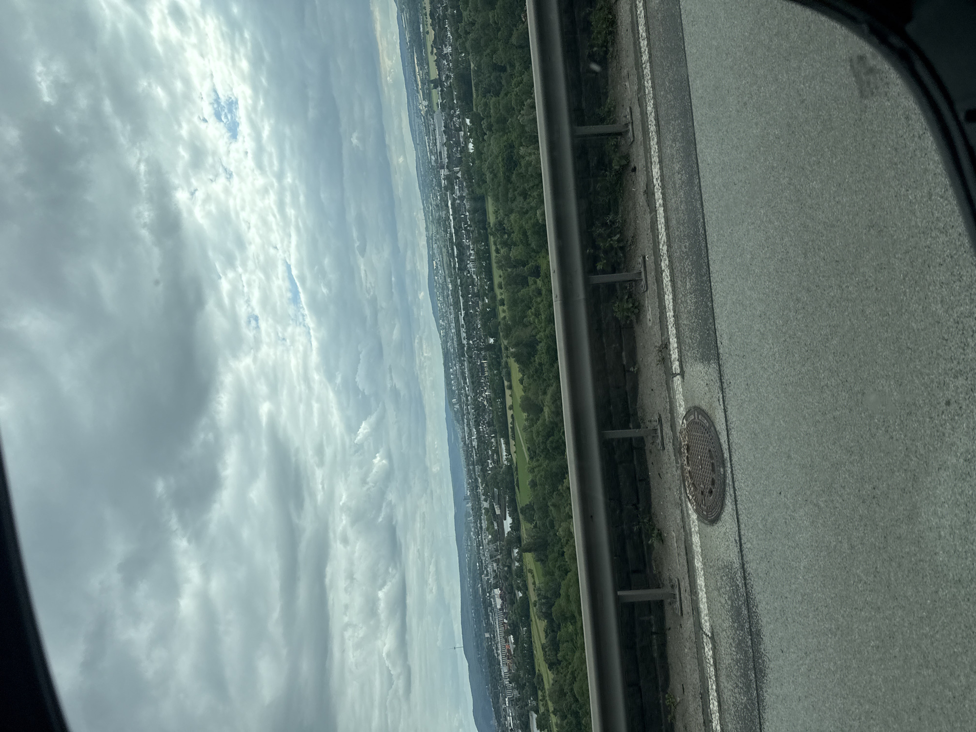 A panoramic view of a city landscape is captured from a high vantage point, likely a bridge or elevated road, looking out over Weitersburg, Germany. The foreground features a gray asphalt road with a white line marking its edge. To the left, a circular metal manhole cover is embedded in the pavement. Beyond the road, a metal guardrail runs horizontally across the image, supported by visible posts. This guardrail separates the road from a grassy embankment with some sparse vegetation.
The middle ground is dominated by a dense, green forest that stretches across the landscape. Interspersed within and beyond the forest are numerous buildings, indicating a populated area, with a river or body of water visible in the distance. The background shows a sprawling city with a mix of residential and industrial structures, extending towards a range of hills under a cloudy, overcast sky. The clouds are thick and gray, with some lighter patches suggesting breaks in the cloud cover, and a faint blue sky is visible in some areas. The overall impression is one of a slightly muted, possibly humid, day with a wide view of urban and natural scenery.