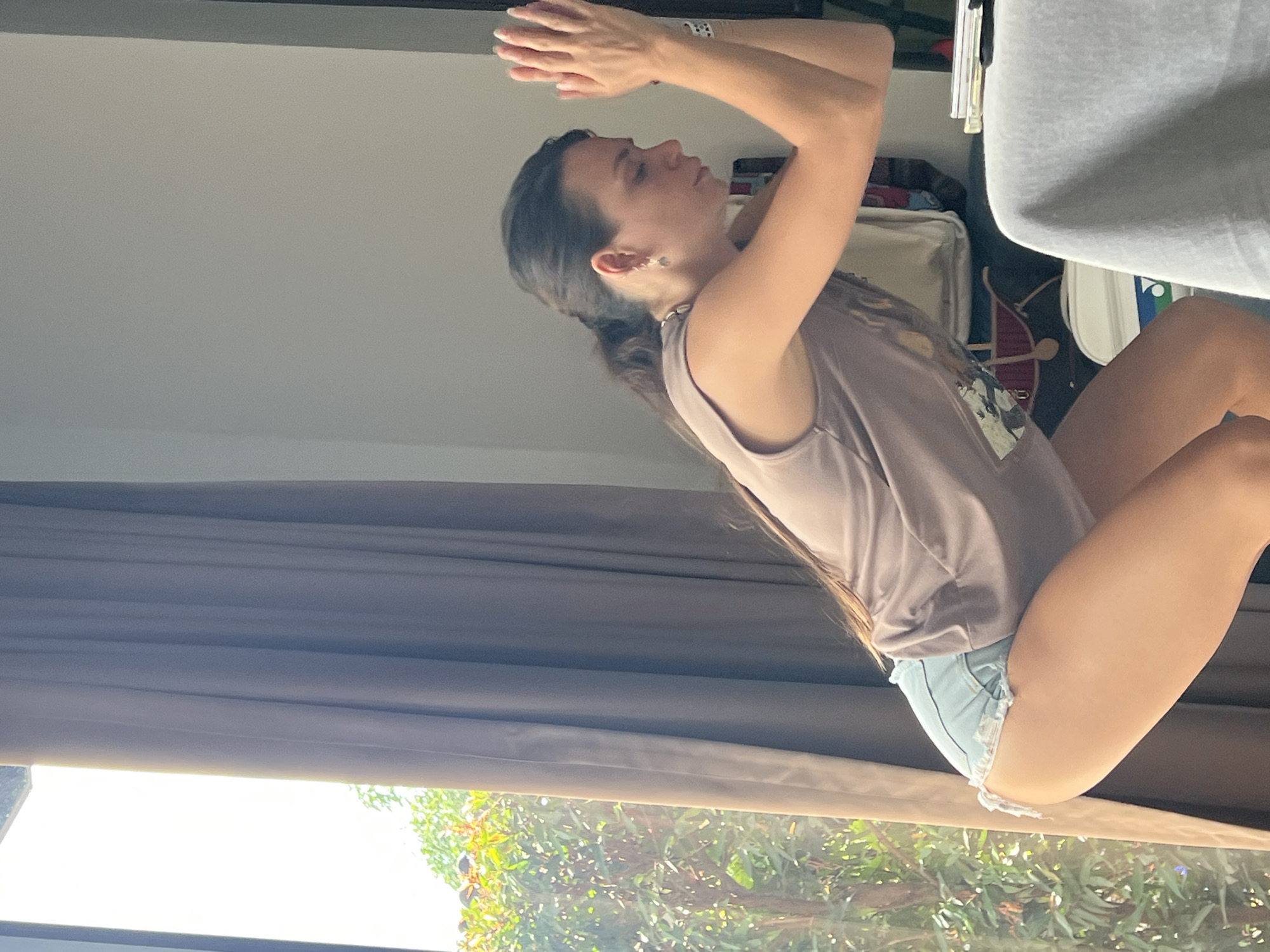 A young woman with dark hair is seen indoors in a room in บ้านบางรักษ์, Thailand. She wears a brown sleeveless shirt and blue denim shorts. Her hands are clasped together above her head. Outside the window are green trees. The room has brown curtains and a gray ceiling. There is a chair and other objects in the background, suggesting this is in a home environment.