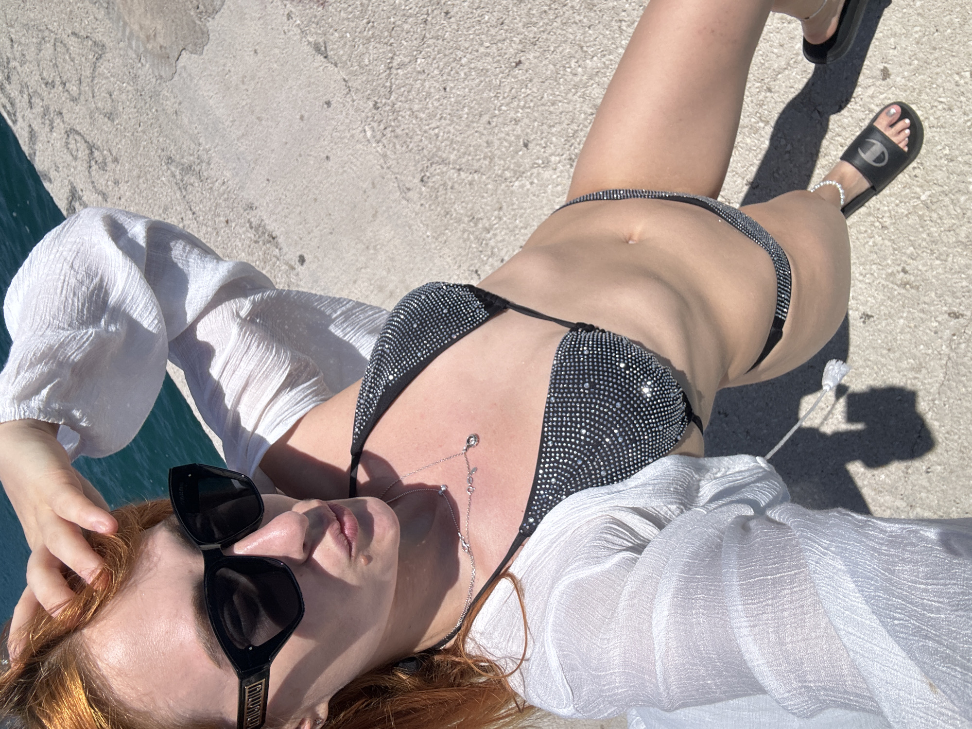 An overhead, high-angle shot captures a person lying relaxed on a light-colored concrete or stone surface under bright, direct sunlight. The setting is outdoors, with a glimpse of blue water visible on the left, suggesting a coastal or waterfront location, consistent with Budva, Montenegro.
The subject, likely female, has reddish-brown hair and is wearing a dark, sparkly bikini top and matching bottoms. A sheer, white, crinkled long-sleeved cover-up is worn over the swimwear, draped around the upper body and arms. Black sunglasses are perched on the face, with "MIU MIU" visible on the arm and a stylized emblem on the temple. A delicate silver necklace with small charms is worn around the neck. On the feet, black slide sandals feature the "Champion" logo. Silver anklets adorn the ankles, and the toenails are painted silver.
The person's pose is casual and leisurely, with one hand resting near their head. Strong, well-defined shadows cast on the ground indicate a clear, sunny day, likely around midday. The overall scene conveys a sense of relaxation and enjoyment of warm weather in a scenic, sunny environment.