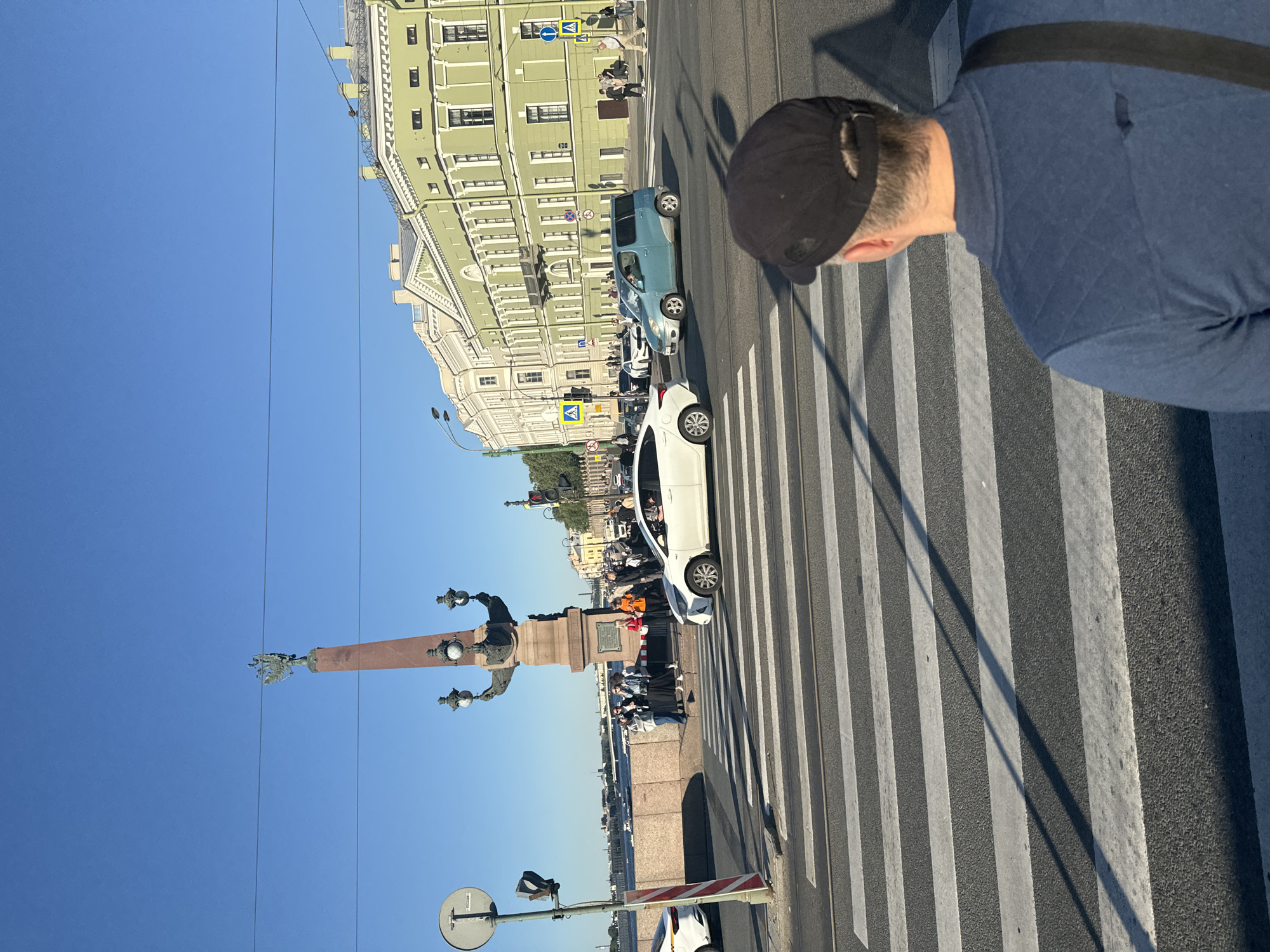 This image depicts a street scene in Saint Petersburg, Russia, likely on a bridge due to the presence of a large stone embankment and a body of water visible in the background. The foreground shows a pedestrian crossing with white stripes on an asphalt road. In the middle ground, several cars are present, including a white sedan and a light blue minivan, navigating the intersection. Pedestrians are visible on the sidewalk and at the crosswalk, some appearing to be tourists. Behind the street, multi-story buildings with distinct architectural styles line the road, painted in light green and white. A prominent monument, an obelisk topped with a double-headed eagle, stands to the left of the frame, alongside street lamps. The sky is clear and blue, indicating a sunny day. The perspective is from behind a person wearing a blue shirt and a black baseball cap, looking towards the street and the cityscape.