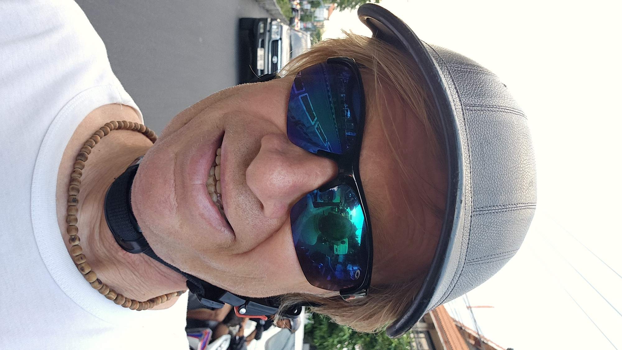 A close-up portrait shows a man wearing sunglasses and a leather helmet. The reflection in his sunglasses shows a cityscape. The man wears a necklace of wooden beads, and the strap of a helmet is visible around his neck. A white t-shirt is partially visible. Cars are parked along a street in the background. The photo was taken in Denpasar, Indonesia.