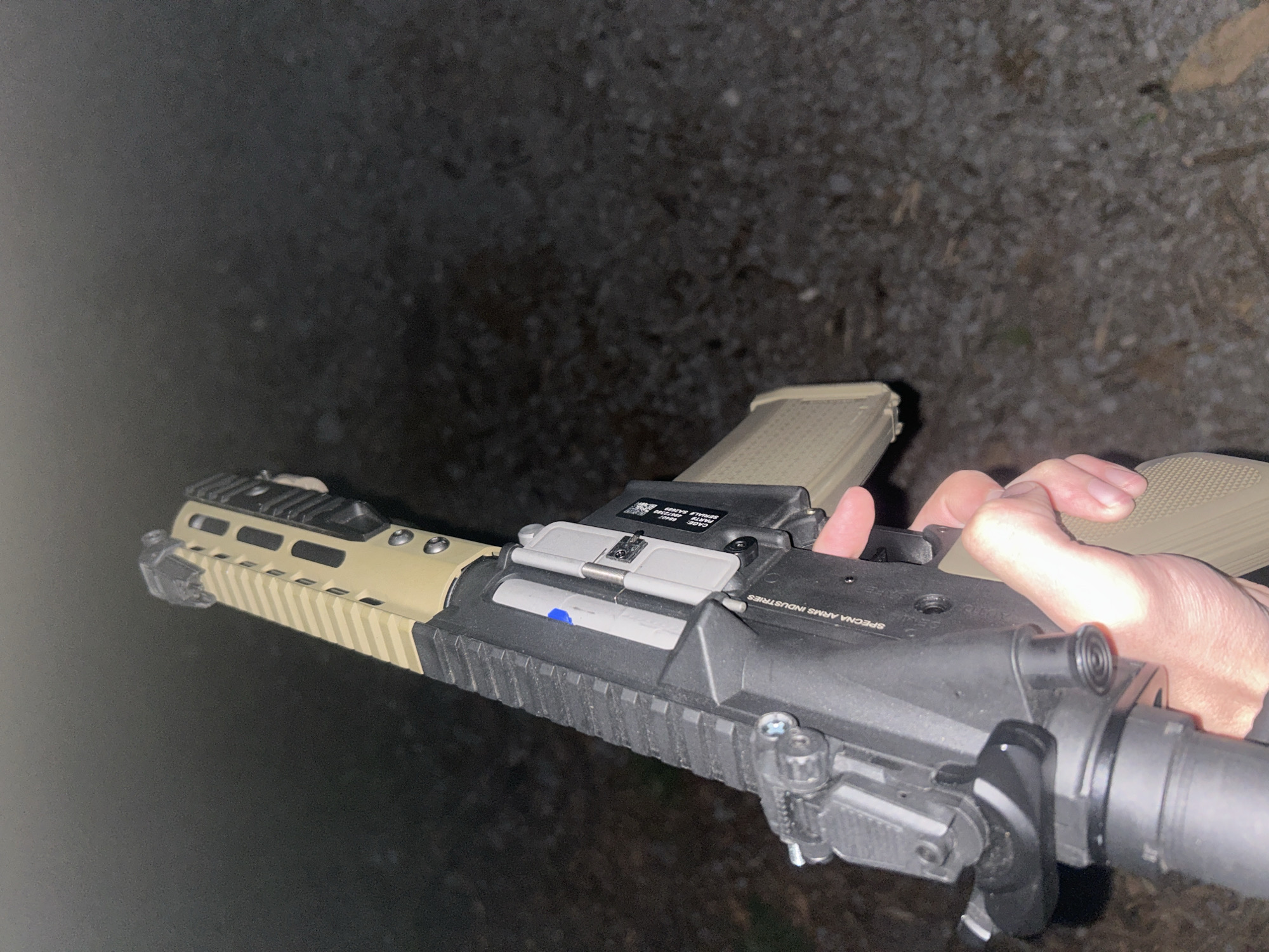 A close-up, low-angle shot shows a person holding an assault rifle, possibly an airsoft replica, outdoors at night. The rifle is mostly black with tan accents on the front grip and magazine. The person's hand is visible near the magazine. The background is dark and out of focus, suggesting a gravel or dirt surface. The rifle has markings that suggest the manufacturer, Specna Arms Industries. The location cannot be determined from the image.