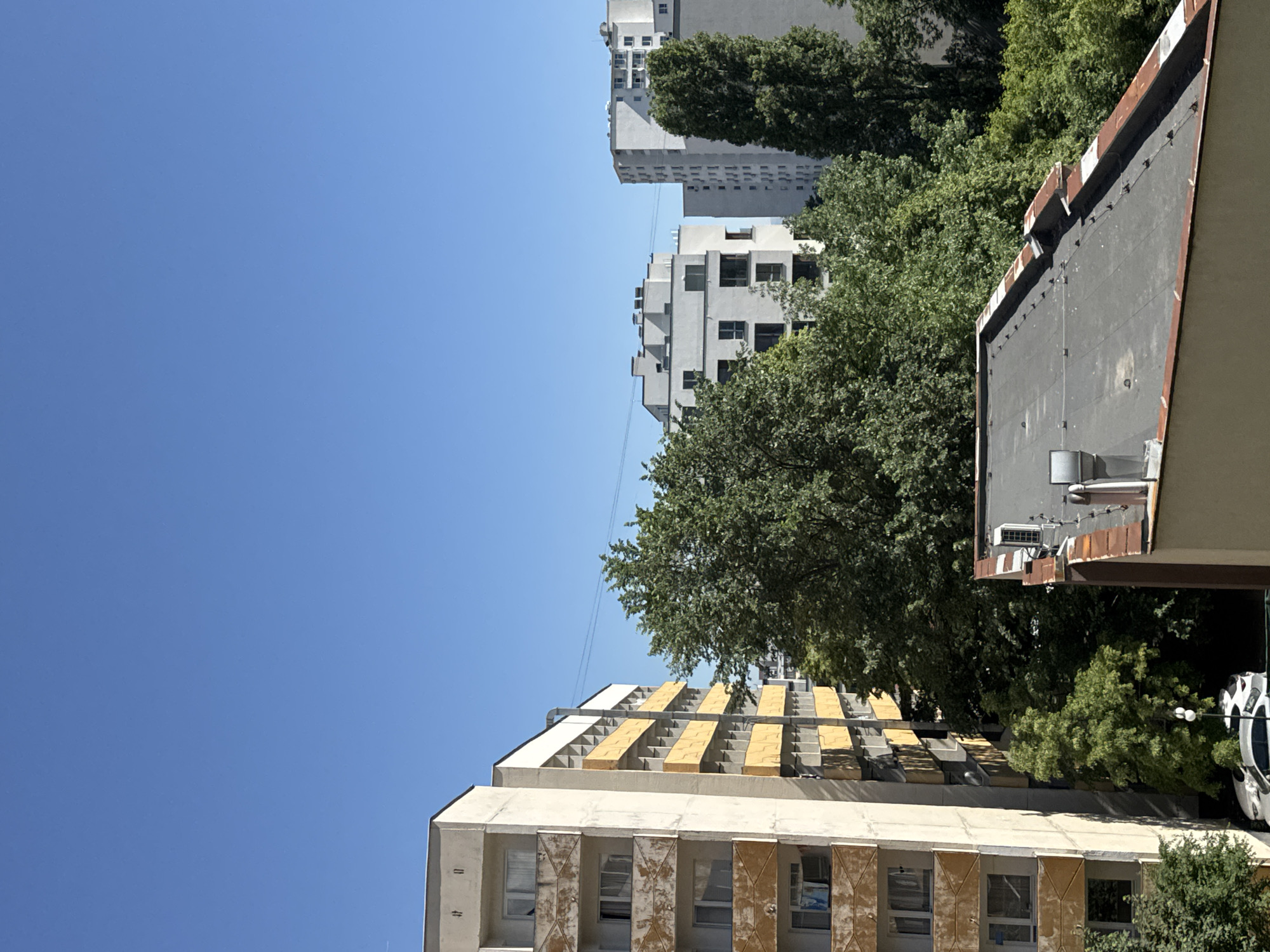 The image shows a view of buildings in Sofia, Bulgaria, under a clear blue sky. There are three buildings visible: the first one has a cream and yellow facade and features a unique geometric design on its upper levels. The second building is white and has a simple, minimalist design with multiple windows, while the third building is mostly hidden behind the trees, but appears tall and grey. Green trees are scattered throughout the view, filling gaps between buildings. In the foreground, a dark-colored building roof is visible. The overall scene conveys an urban landscape on a sunny day.