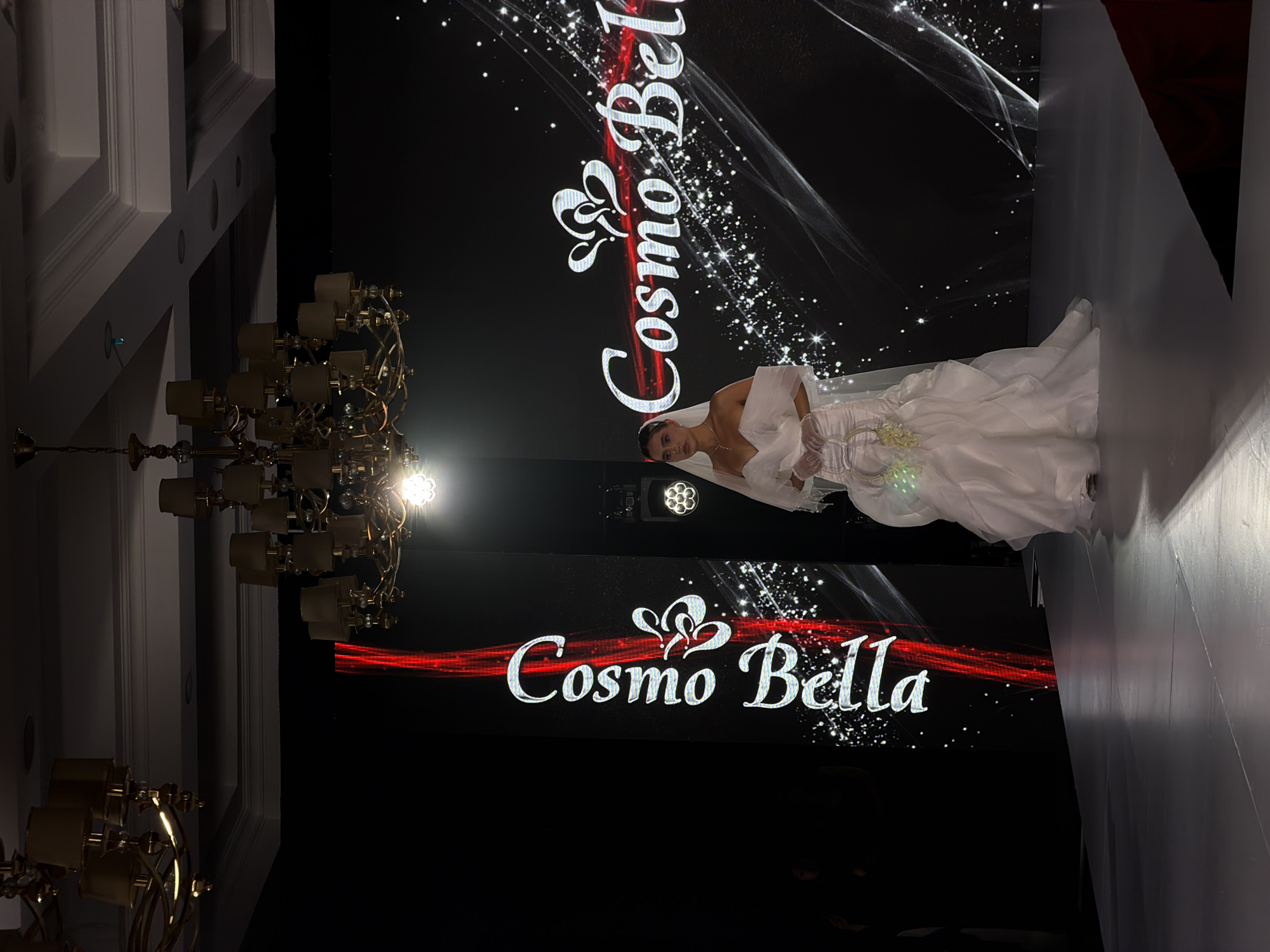 A woman in a white wedding dress and veil stands on a white runway. Behind her is a large black screen that displays the words "Cosmo Bella" in a white, cursive font with red accents and decorative flourishes. The screen also has a black background with white light streaks and dots. A large chandelier hangs to the left of the screen, and there are stage lights visible above the model. The scene appears to be taking place at a fashion show in the city of Unknown, Unknown.