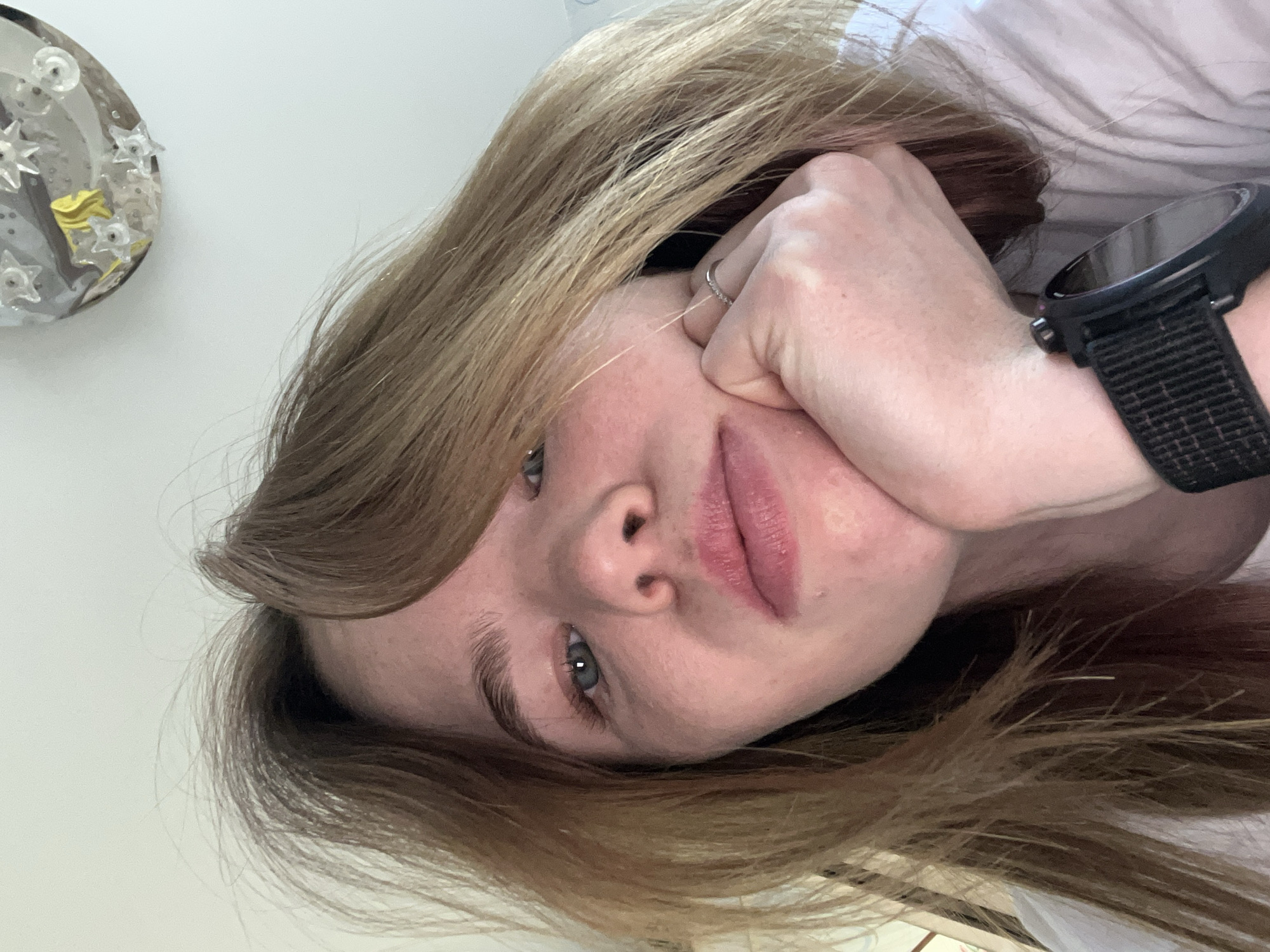 A close-up, indoor, slightly angled portrait features a woman with light blonde and brown hair. She is resting her face on her fist, which has a thin ring on the ring finger. She wears a black smartwatch on her wrist. The background consists of a white wall with a small, decorative, round object with stars on it attached, along with a white surface, possibly a bed. The location and the city of Chaikovsky, Russia, cannot be determined from the image.