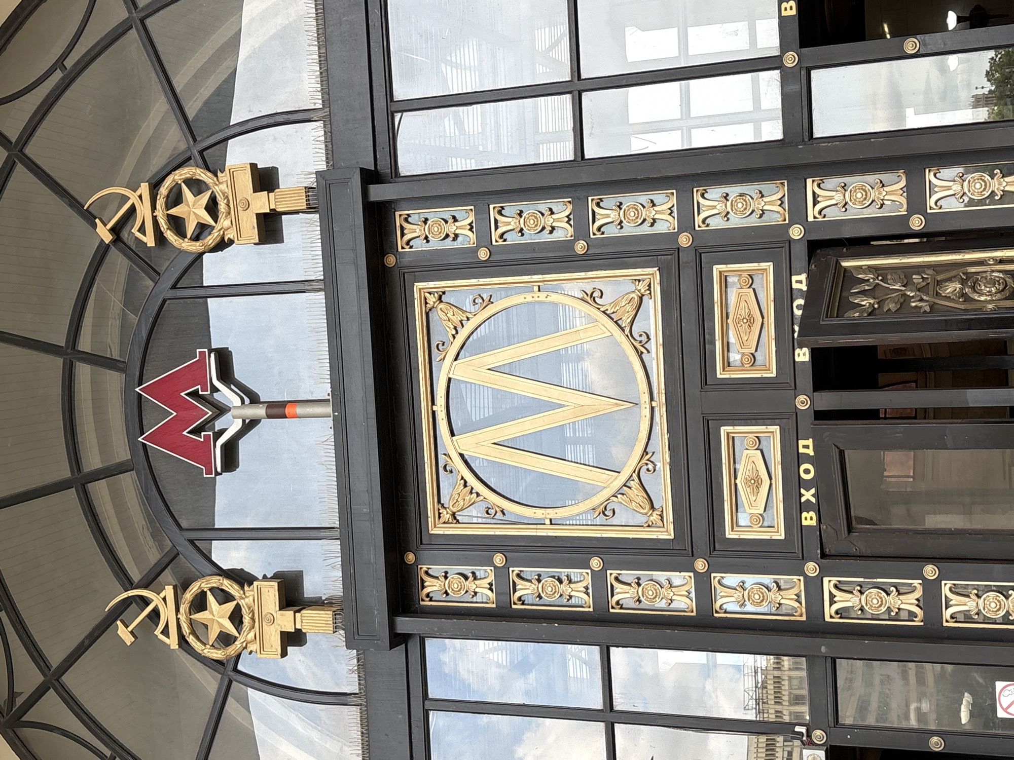 The image shows the entrance to a Moscow Metro station in Russia. The entrance features a large "M" symbol for the metro, along with hammer and sickle and a star with wreaths around it. The entrance is a black frame with gold-colored decorations. The sign that appears to say, "Вход," is in Russian and means "Entrance." The design reflects Soviet-era architecture and aesthetics. The entrance is framed by windows and ornate metalwork. The windows reflect the city surroundings.