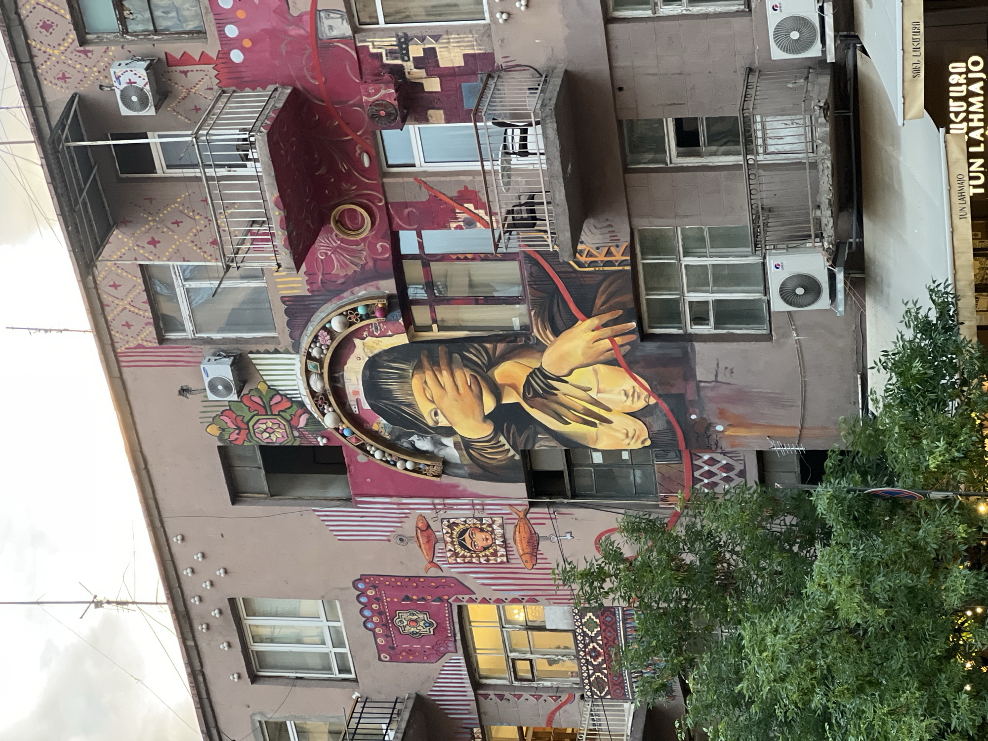 A low-angle, full outdoor shot depicts the exterior facade of a building in Yerevan, Armenia, showcasing a variety of window types, balconies, and a complex mural covering a significant portion of the building's exterior.
The building is painted a neutral tan color, serving as a background for the vibrant mural. The mural features a diverse array of images and designs, including a large painting of a woman covering her eyes, surrounded by an embellished border. Other parts of the mural include designs inspired by Armenian carpets, floral motifs, geometric patterns, and figures of fish. The mural elements incorporate a range of colors, including shades of red, gold, brown, and touches of turquoise.
The building has multiple windows of varying sizes and styles, some reflecting the ambient daylight. Several balconies with simple metal railings are also integrated into the building’s design. Modern air conditioning units are mounted on the facade.
On the right side of the image, a glimpse of a storefront sign is visible.
The scene is framed by the foliage of trees in the lower right corner, with additional cables or wires extending across the sky towards the upper left corner.