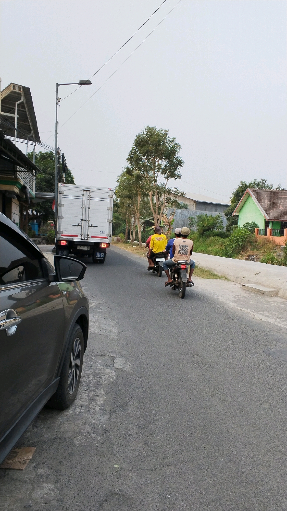 The image depicts a road scene in Banjaragung, Indonesia, with a dark-colored car in the foreground on the left. On the road, a white truck is moving away from the viewer, followed by two motorcycles. The first motorcycle has a person in a yellow shirt. The second motorcycle carries two people, one wearing a blue shirt and the other in a light-colored shirt with a QR code design. Trees and buildings are visible on the sides of the road, with a light-colored sky overhead.