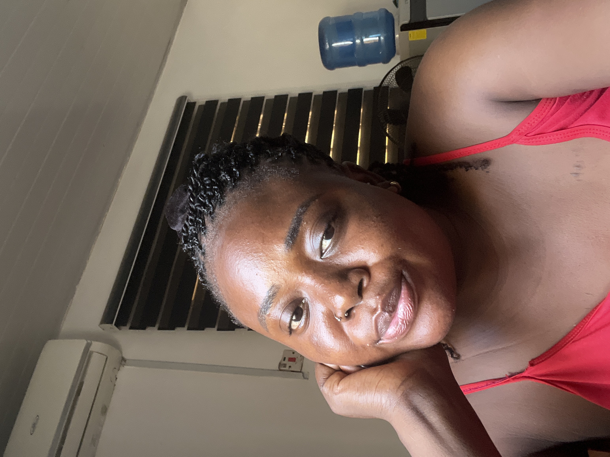 This media file is a close-up, Dutch-angle selfie of a Black woman. She is resting her chin on her hand and looking towards the camera with a gentle smile. She has dark, curly hair braided and a nose ring. She is wearing a red top.
In the background, a black fan is visible behind horizontal blinds. Above the blinds, a blue water jug is mounted on a white surface. To the left of the frame, a white air conditioning unit is mounted on a wall.
The lighting suggests it is daytime, possibly indoors. The presence of the air conditioning unit and the water dispenser hints at a warm climate, consistent with the provided location context of Abuja, Nigeria. The overall mood is relaxed and informal. No visible text is present in the image.