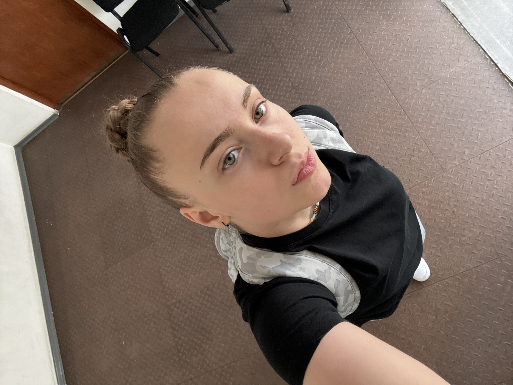 A young woman stands indoors, looking up into the camera. She has fair skin, green eyes, and her hair is pulled back into a bun. She is wearing a black t-shirt and a light gray patterned backpack. The floor is brown with a textured surface. In the background are black chairs and a wooden door. There is a white wall or window ledge in the upper right corner. The location cannot be determined as Sofia, Bulgaria based on the image alone. No specific objects or interactions suggest its location.