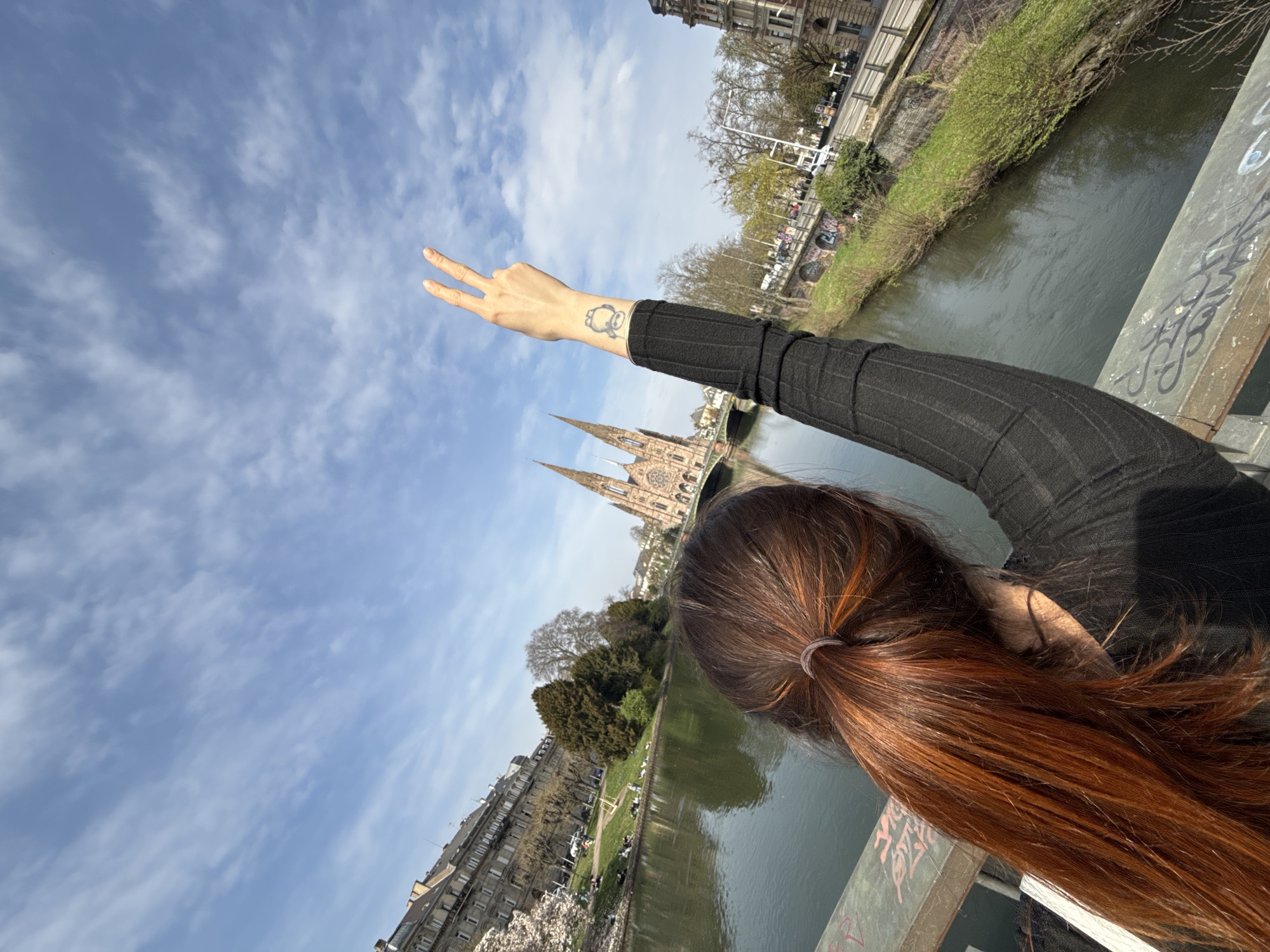 The image depicts a daytime scene in Strasbourg, France, observed from a bridge spanning the Ill River, with Église Saint-Paul (Saint-Paul's Church) as the central distant landmark. In the foreground, a person is seen from behind, their head tilted slightly to the right. The person has long, reddish-brown hair tied in a ponytail and wears a black, long-sleeved ribbed top. Their right arm is raised, with the hand forming a "peace" sign (index and middle fingers extended). A circular tattoo is visible on the inner wrist of the raised arm. The person is leaning against a gray-green metal bridge railing, which is marked with black and red graffiti.
Beyond the railing, the Ill River flows. The river's left bank features a grassy park area where several individuals are seated or reclining on the ground. Behind the park, a long, multi-story building with classical architecture is visible, along with trees, some of which are in bloom with white flowers. On the river's right bank, a paved road with vehicles and streetlights runs parallel to the water, bordered by a stone wall also bearing graffiti and sparse vegetation. Further down the right bank, additional multi-story buildings with traditional European architecture are present.
Dominating the midground-background is Église Saint-Paul, a large Neo-Gothic church constructed from reddish-brown stone, featuring two tall, pointed spires. The church is situated on an island formed by the Ill River, with a bridge visible leading to it. The sky above is clear blue with scattered, wispy white clouds, indicating fair weather conditions and bright natural illumination.