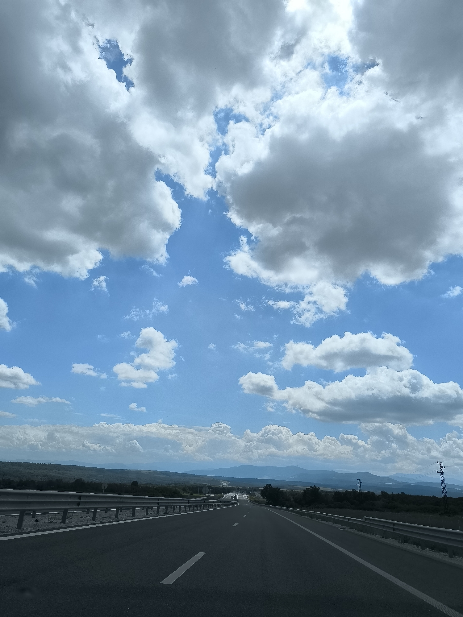 The image displays a landscape scene captured from a vehicle on a multi-lane asphalt highway in Valchek, Bulgaria. The road, delineated by white solid and dashed lane markings, stretches into the distance, with a metal guardrail running parallel on both sides. To the left of the road, a strip of ground is bordered by a guardrail, followed by a vegetated area. To the right, a similar guardrail separates the road from an unpaved shoulder and dense green vegetation, including trees and shrubs. In the mid-ground on the right, multiple dark-colored power line towers are visible against the distant landscape. The background features rolling hills and mountains extending across the horizon, appearing bluish due to atmospheric perspective. The sky, occupying approximately two-thirds of the upper frame, is predominantly bright blue, punctuated by numerous large, fluffy cumulus clouds ranging from brilliant white to light grey. Patches of clear blue sky are evident between the cloud formations. No individuals or other vehicles are explicitly depicted.
