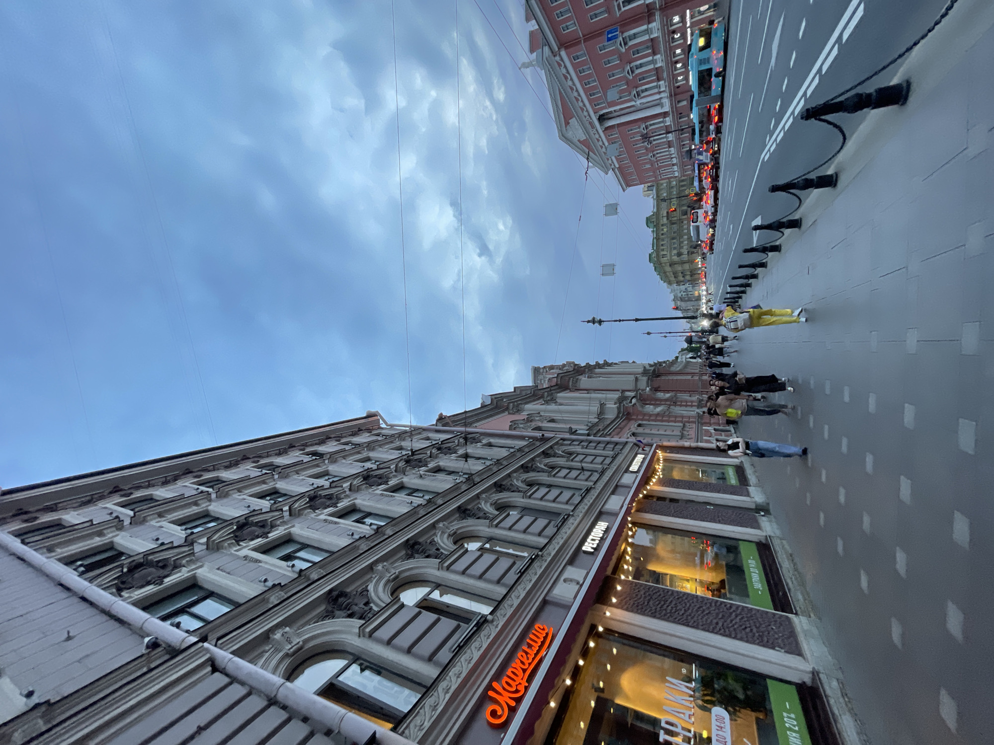 An eye-level, wide-angle outdoor shot depicts a street scene in Saint Petersburg, Russia, at dusk. The image is dominated by the imposing architecture of buildings lining the left side of the street, with ornate facades and rows of arched windows. A prominent neon sign in Russian, which translates to "Marshmeloe," is visible on one of the buildings, indicating a retail establishment. Pedestrians walk along the sidewalk, their figures blurred by motion.
Across the street, traffic moves in a single direction, including buses, cars, and a tram. The road is well-lit, and streetlights cast a soft glow. The sky is overcast with dramatic, dark clouds, suggesting an impending change in weather. The overall atmosphere is one of urban life transitioning into evening, with a mix of historical architecture and modern transportation.
The specific location appears to be Nevsky Prospekt, a major thoroughfare in Saint Petersburg, known for its grand buildings, shops, and bustling atmosphere. The style of the buildings, the presence of a tram, and the general urban character are consistent with this iconic street.