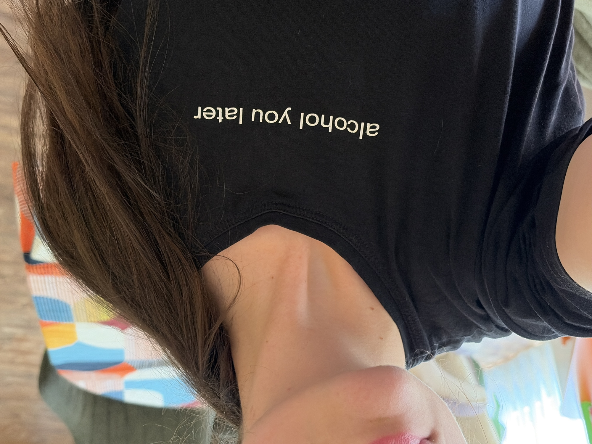 A close-up, slightly overhead shot captures the upper body of a person, primarily focusing on their black t-shirt and flowing brown hair. The person's long, dark brown hair cascades down their back and shoulder, partially obscuring the background. They are wearing a black short-sleeved t-shirt with white text emblazoned across the front. The visible text clearly reads "alcohol you later."
The image also shows a portion of the person's neck, shoulder, and a hint of their chin and lips, which appear to have a pink tint. In the background, behind the person's hair and shoulder, there's a glimpse of a colorful, patterned object, possibly a blanket or fabric, with abstract geometric shapes in shades of orange, blue, yellow, and white. The lighting suggests an indoor setting during daylight hours. No specific activity is depicted other than wearing the t-shirt. The person's facial expression or specific location cues are not discernible.