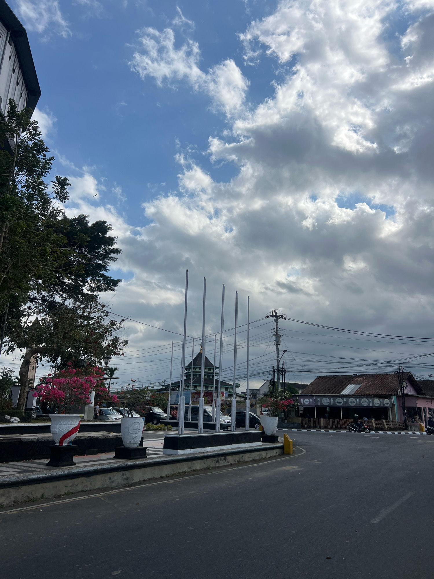 The image depicts a street scene in Magelang, Indonesia, on a partly cloudy day. In the foreground, a paved road curves to the right. A raised concrete median runs along the left side of the road, adorned with two large white planters containing vibrant pink flowers and greenery. Behind the median, several tall, slender flagpoles stand in a row.
To the left, a portion of a modern building with a dark roof and white walls is visible, partially obscured by lush green trees. Further back, a building with a green roof and ornate architecture is also present. The middle ground features more buildings with terracotta roofs and various shops lining the street. Electrical wires and a utility pole are prominent overhead, indicating an urban setting. A few cars and motorcycles are present on the road and sidewalk, with some people seen in the distance. The sky is a mix of blue with dramatic, gray clouds.