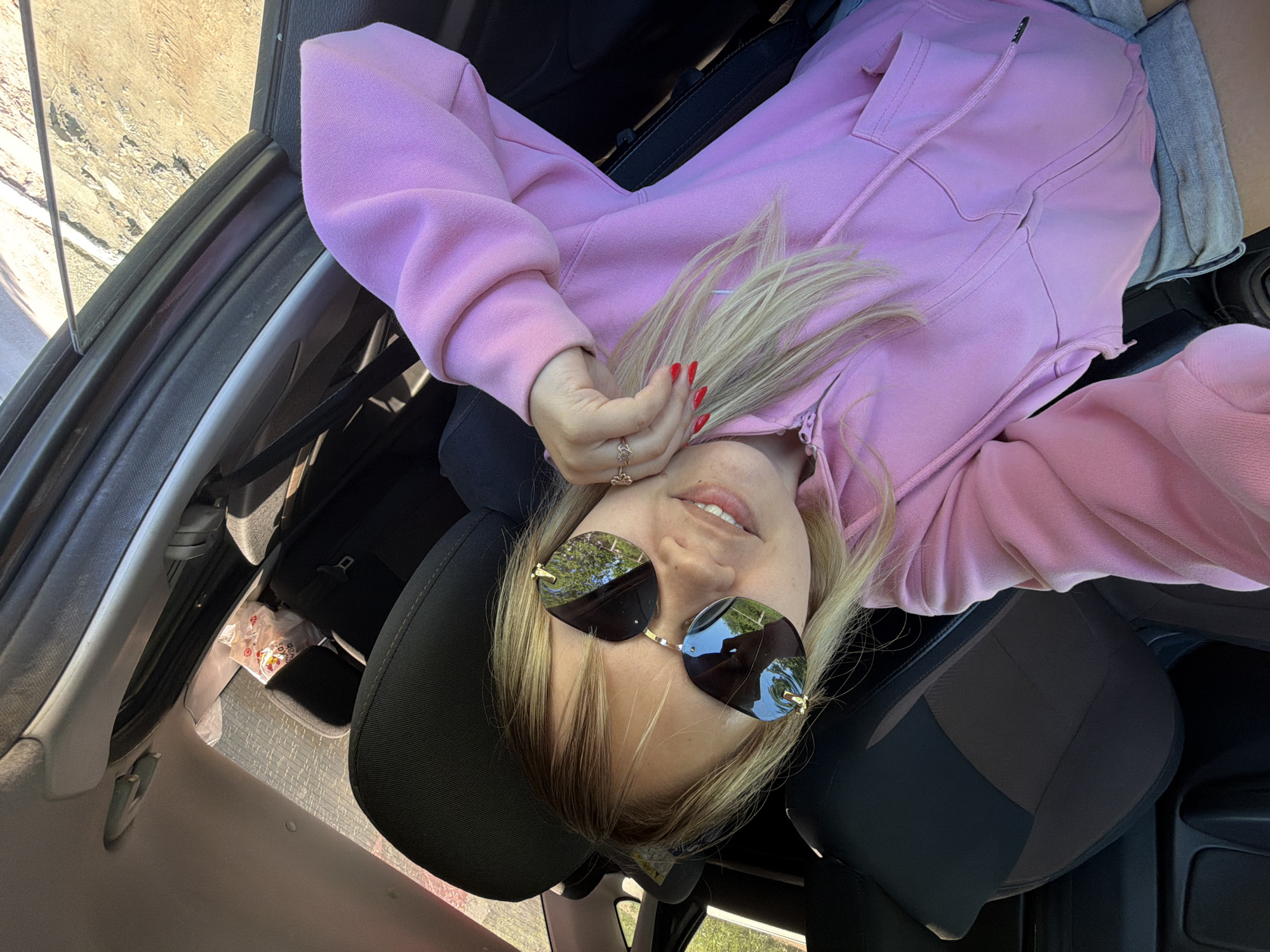 A woman with blonde hair is reclining in the driver's seat of a car. She is wearing a pink hoodie and sunglasses. Her red painted fingernails are visible on her left hand. The reflection in her sunglasses shows trees and blue sky, suggesting a park or outdoor setting. A fast food paper bag is visible in the car's interior. The photo might have been taken in Chaikovsky, Russia.