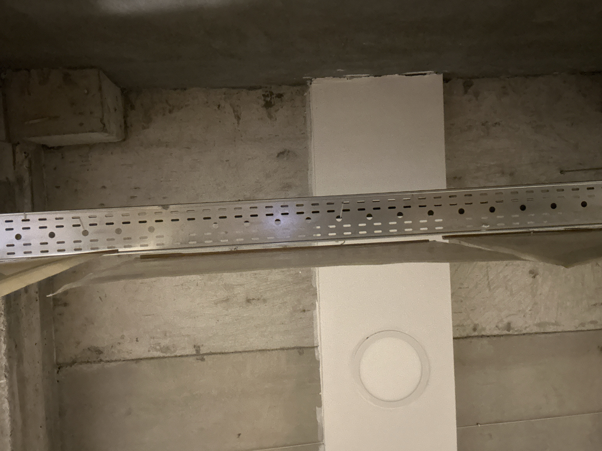 The image depicts an interior, likely in a basement or utility space, given the exposed concrete structure. The scene is characterized by unfinished walls and ceilings, with a metallic cable tray running horizontally across the frame. This tray is perforated with a repeating pattern of slots and circular holes, suggesting its use for routing electrical or data cables. A vertical white rectangular element, possibly a duct or pipe enclosure, is visible, featuring a circular recessed light fixture. The lighting in the space is dim, casting shadows and creating a sense of depth. The environment appears industrial or utilitarian, lacking any decorative elements or signs of regular habitation. The location and city are unknown.