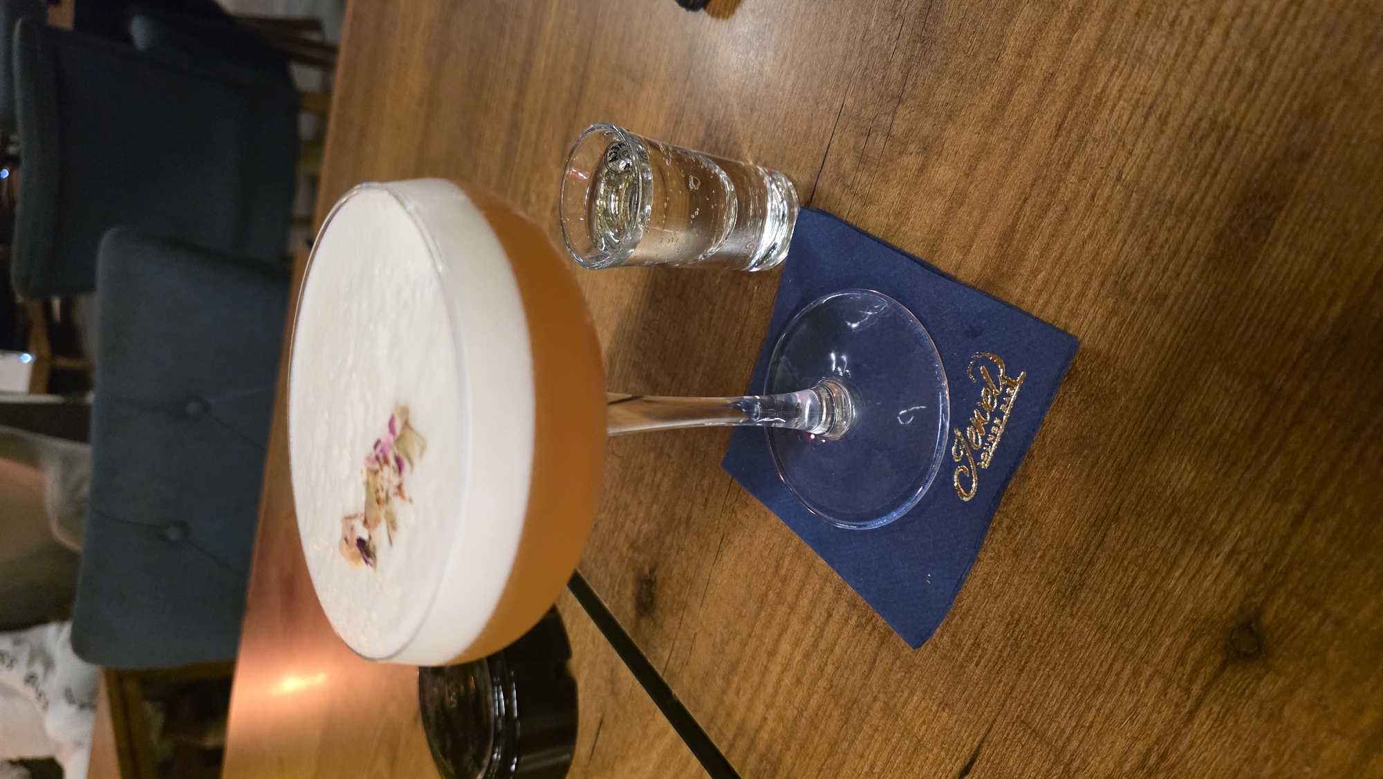 An overhead shot captures a cocktail and a shot glass on a wooden table, likely in an indoor setting like a bar or restaurant during the evening. The image is oriented with the table appearing to slant downwards from left to right.
The prominent subject is a cocktail served in a clear coupe glass, positioned in the lower-left to mid-left of the frame. The drink has a vibrant amber or orange hue at the bottom, topped with a thick layer of creamy white foam. This foam is delicately garnished with small, dried flower petals in shades of pink and beige, scattered across its surface.
To the right of the cocktail, a small shot glass filled with a clear liquid, possibly water or a spirit, rests on the wooden surface. The shot glass is situated on a dark blue napkin.
The dark blue napkin, which serves as a coaster for the cocktail glass's stem, features a distinctive gold-colored logo or text. The text is in a stylized script font and appears to read "Jewel".
The table itself is made of wood, showcasing a rich brown color with visible grain patterns. In the blurred background, there are hints of dark upholstered chairs and soft ambient lighting, further suggesting a cozy, indoor establishment. The overall atmosphere implies a relaxed and social occasion.