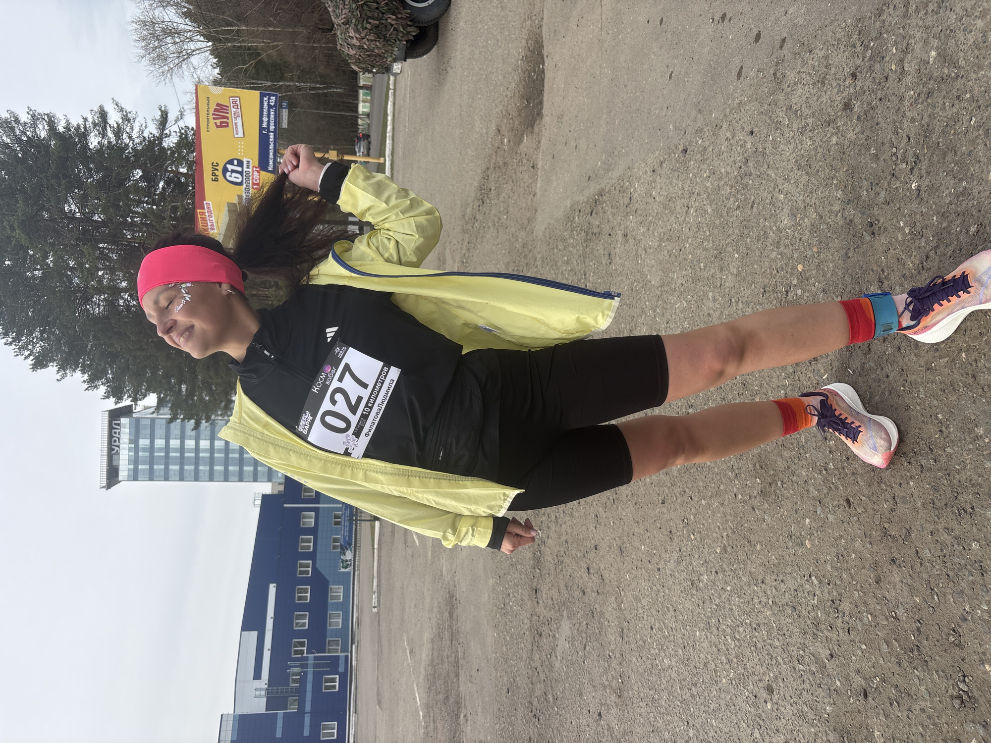 A woman with a pink headband, wearing a black running outfit with a yellow jacket and running shoes, is shown participating in the Kosm Zabeg (Cosmos Run) in Neftekamsk, Russia. Her race number is 027. The event is a 10-kilometer race for Filatova Lyudmila. In the background, there is a building with "URAL" written on it, a blue building with white windows, trees, and a construction advertisement. The ad includes the text: "AKTSIA VYGODNO" (promotion profitably), "BRUS," "STROITELNYJ BOOM" (Construction Boom), and "G. NEFTEKAMSK, KOMSOMOLSKIY PROSPEKT 43d." The woman is standing on asphalt.