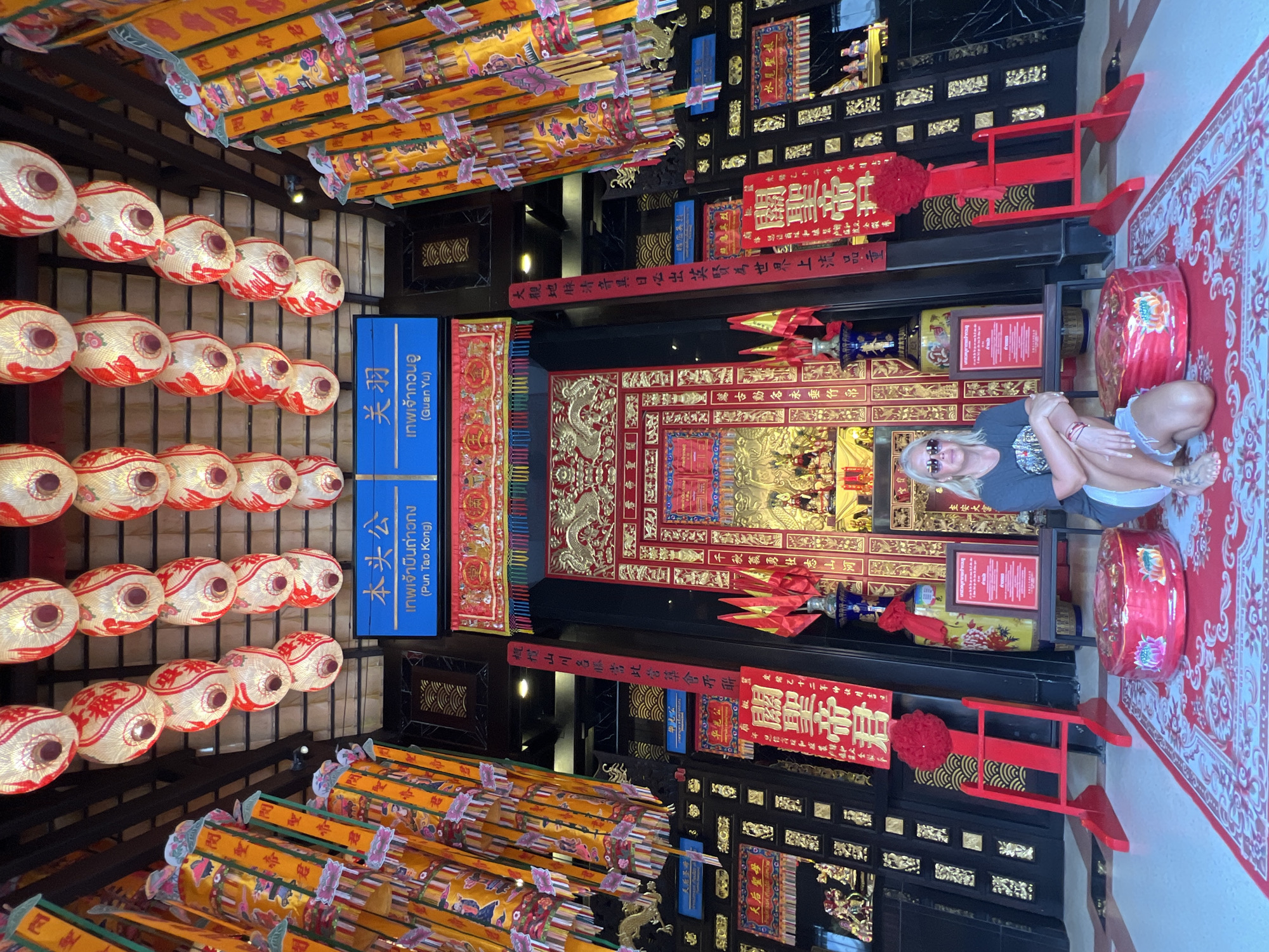 A medium shot captures a person in a seated, relaxed pose within a vibrantly decorated setting. The person, with blonde hair and sunglasses, is wearing a dark t-shirt and shorts, with tattoos visible on their feet. They are positioned in front of a richly adorned shrine-like structure, featuring intricate gold carvings, red banners with Chinese calligraphy, and decorative elements. The scene appears to be indoors, possibly a temple or a cultural establishment, given the ornate decorations and the presence of lanterns and religious iconography.
Rows of round, white lanterns with red and gold accents hang from above, adding to the festive and traditional atmosphere. In the background, a large red drum with decorative patterns sits on a red carpet. Blue signs with Thai and Chinese text are visible, with one clearly displaying "เทพเจ้ากวนอู (Guan Yu)", indicating a dedication to the deity Guan Yu. The overall impression is one of cultural immersion, likely in a location with a significant Chinese influence, such as Baan Hua Thanon, Thailand. The lighting suggests it could be daytime, though the interior setting limits definitive conclusions about the time of day or weather. The person's relaxed posture and the ambient decorations create a sense of tranquility and engagement with the surroundings.
--- OCR Start ---
σ
本头公
เทพเจ้าบินถ่าวกง
(Pun Tao Kong)
关羽
เทพเจ้ากวนอู
(Guan Yu)
君
水尾聖娘
關聖帝丹
光二十二年号我建吉
大觀地候清音具日必出英賢為世界上流品重
萬古動名永垂竹串
主安大家
千秋義勇壮志山河
概慣山川各縣當此營築會否聯
定豬乙十二年的让月吉
關聖帝君
天后聖母
67
--- OCR End ---