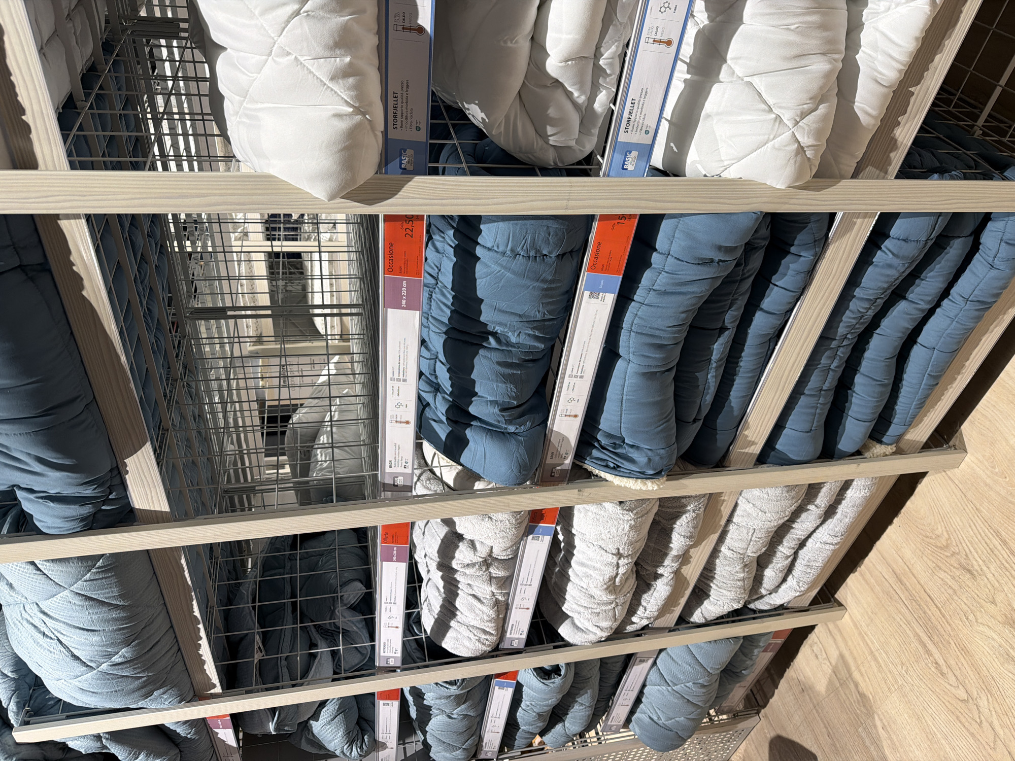 The image depicts a shelf unit in a retail setting, possibly a department store in Ragusa, Italy. The shelves contain stacks of bedding products, including quilts and comforters, displayed in various colors: white, blue, and gray. Each item is packaged with a tag indicating its dimensions, price, and the "BASIC" brand name. Labels include descriptions of product names and material composition, indicating information such as "Occasione" and "STORFJELLET," along with the size of the item (e.g., "240 x 220 cm"). The shelves are made of light-colored wood and wire, creating a grid-like structure. The floor is wood-colored. There are no people visible.