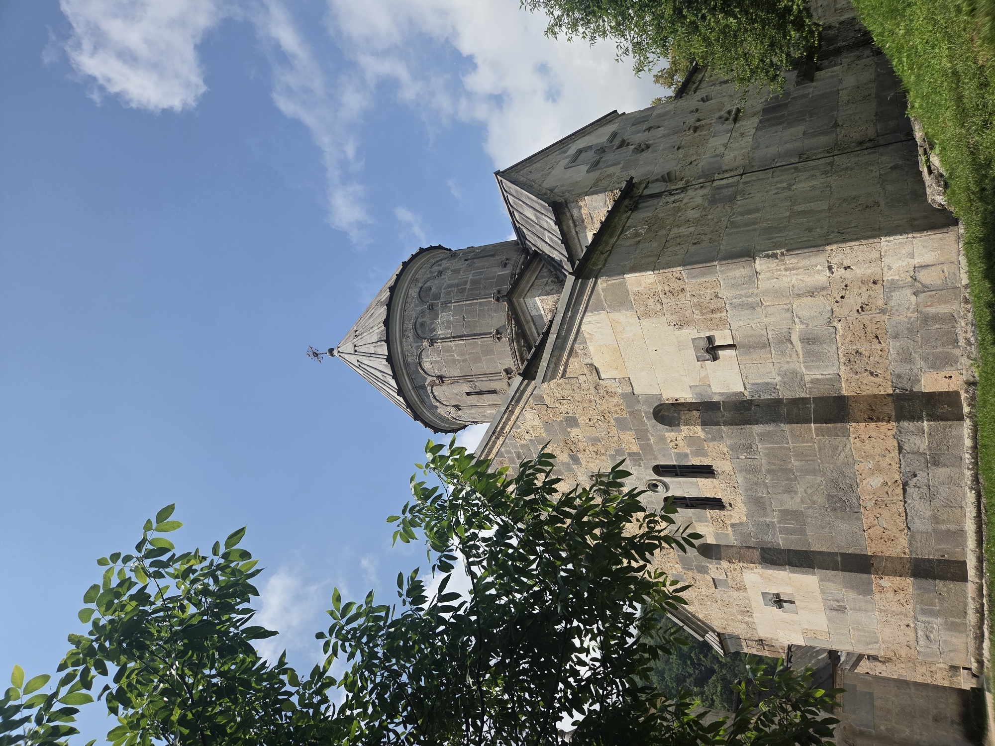 The image shows the exterior of a stone church, likely the Teghut Monastery in Teghut, Armenia, viewed from a low angle. The church is constructed from large, rough-hewn stones in shades of beige and gray. It features a domed roof with a cross at its apex, and a steeply pitched roof section with a metal covering. The building has arched windows and carved cross motifs on its walls. Lush green trees with dark leaves frame the left side of the image, partially obscuring the church and the bright blue sky above, which is dotted with fluffy white clouds. The ground in front of the church is covered with green grass.