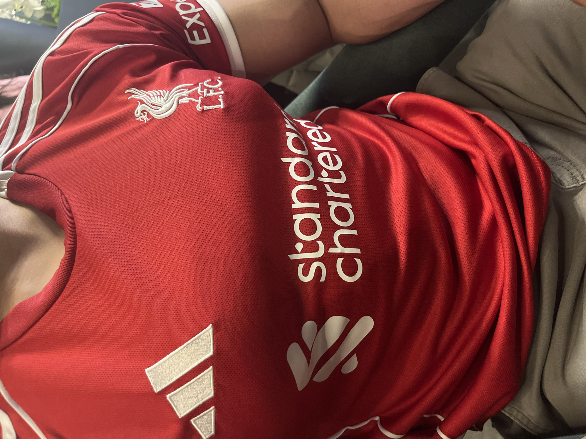 A person is wearing a red Liverpool Football Club soccer jersey with white Adidas stripes on the shoulders, a Liverbird logo on the left chest, and the "Standard Chartered" sponsor logo vertically aligned on the right side of the chest. The location is not discernible from the image and therefore no location information can be provided.