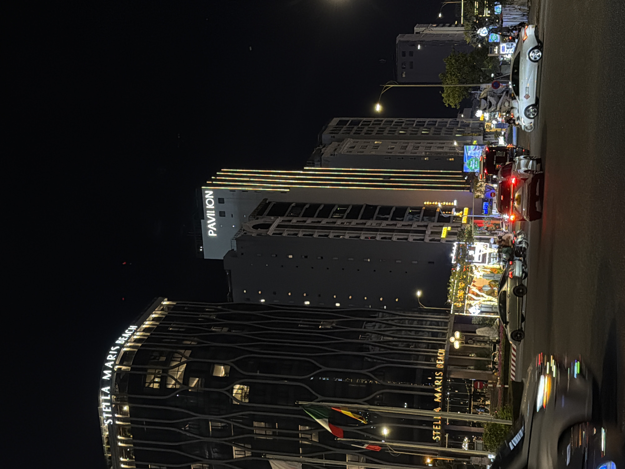 This is a nighttime, eye-level shot taken from a vehicle moving through a city street in Da Nang, Vietnam. The scene is dominated by modern architecture illuminated by streetlights and building lights. On the left, a large, dark building with a distinctive wavy facade prominently displays the illuminated text "STELLA MARIS BEACH." Further down the street, another tall, grey building features the illuminated sign "PAVILION."
The street is busy with traffic, including cars and motorcycles, creating reflections on the wet asphalt, suggesting it may have recently rained or is humid. The atmosphere is lively and urban, with numerous lights from businesses and street lamps creating a vibrant, nocturnal cityscape. The time of day is clearly night, with the sky being dark. The overall impression is one of a bustling city at night, showcasing its commercial and architectural features.