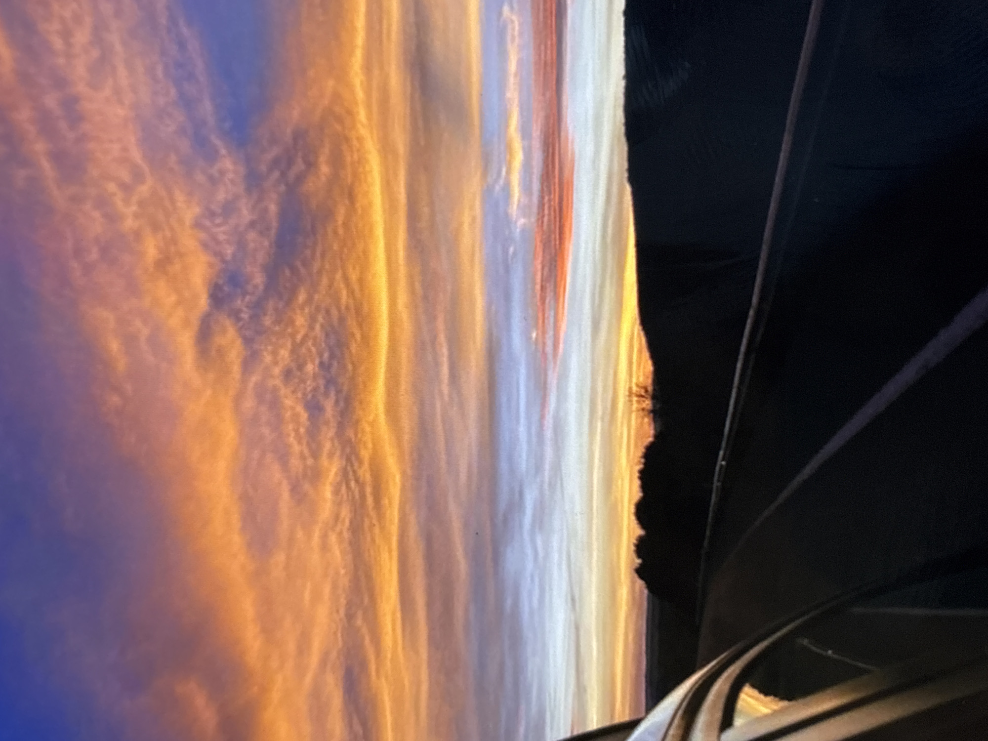 The image depicts a vivid sunset with orange and blue hues dominating the sky, viewed through the interior of what appears to be a vehicle. The vehicle's frame and interior elements are visible on the right side and bottom of the frame, providing a foreground to the sky's dramatic display. The specific location within the city of Belopoptsi, Bulgaria, cannot be determined from the visual information provided.