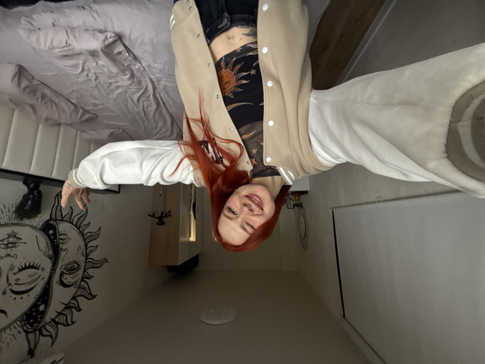 The image depicts a residential bedroom scene, understood to be located in Rostov-on-Don, Russia. A single female subject, appearing in her 20s or early 30s, occupies the foreground. She has long red hair, a light complexion, a septum nose ring, and a visible navel piercing. Her attire consists of a beige bomber jacket with white sleeves and trim, worn over a black mesh crop top featuring a central sun motif. Dark trousers or jeans are visible below her midriff. The subject's arms are extended forward, indicating she is holding a device, presumably a smartphone, to capture a self-portrait. She is smiling broadly with her eyes partially closed, displaying a visible dimple on her left cheek.
The background features an unmade bed with light grey bedding and a white, panel-style upholstered headboard. A large black and white tapestry, depicting an intertwined sun and moon with celestial elements, adorns the wall directly above the headboard. To the right of the bed, a dark, minimalist nightstand supports a small, dark, sculpted bust. On the wall to the left, a white roller blind or projector screen is fully extended. A light-colored wardrobe or cabinet is partially visible in the background on the right. A small, oval, ornate mirror is affixed to the left wall above a white surface. A circular ceiling light fixture provides overhead illumination. The room is generally well-lit with ambient light.
