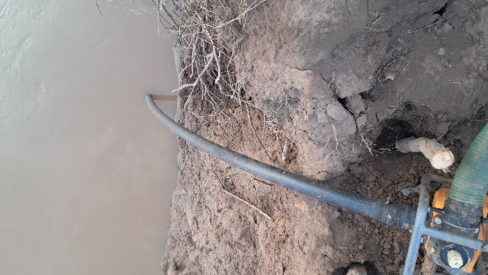 A forensic examination of the scene in Walu, Kenya, reveals a water pumping operation situated on a steep, eroded riverbank. The upper portion of the image displays a body of light brown, turbid water exhibiting minor surface ripples, indicative of a river or large stream. Extending from the lower left frame and curving into the muddy water is a large, black, flexible corrugated pipe, approximately 6-8 inches in diameter, appearing to function as an intake. A light-colored wooden stake is positioned near where the pipe enters the water.
The riverbank itself consists of dark brown, loose, and crumbly soil, showing signs of erosion and containing scattered dry branches and plant debris. In the lower left quadrant, partially visible, is the base of a mechanical pump unit, characterized by a dark metal frame and visible orange components. Connected to this unit is a green corrugated hose, extending out of the frame to the lower left. To the right of the pump, a cut wooden stump or thick root is embedded in the soil, adjacent to a small depression in the bank. No individuals are present in the frame.