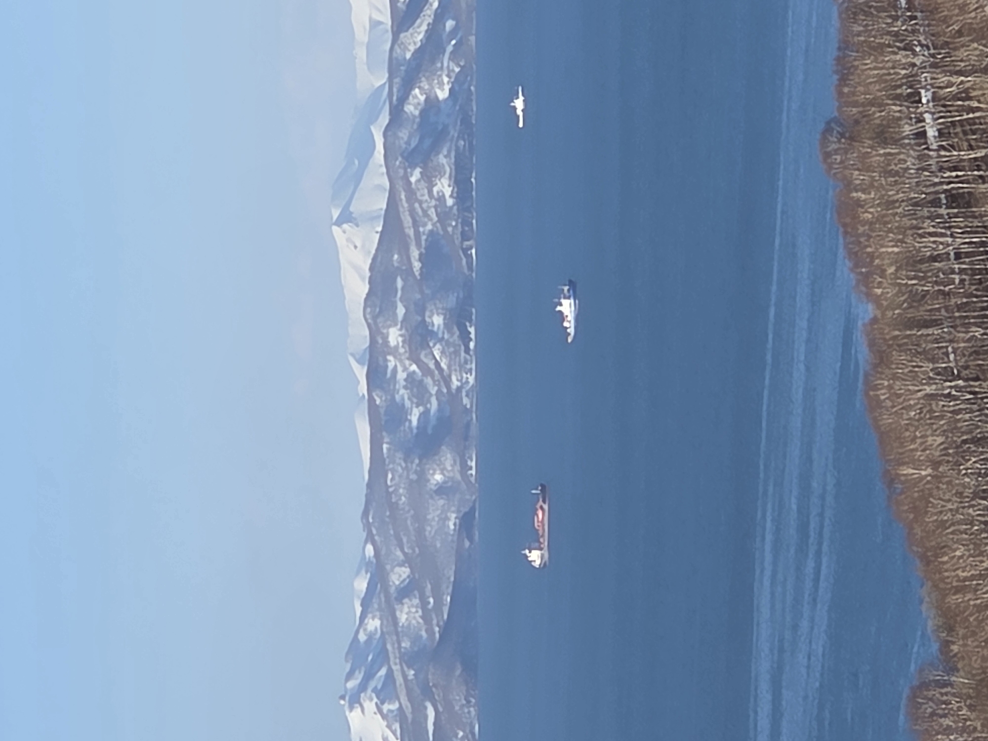 The image shows a body of water, likely a sea or large lake, with several ships or boats visible on the water's surface. Mountains are visible in the background, partially covered in snow, indicating a cold or mountainous region. The foreground features a shoreline with vegetation, potentially trees or shrubs. The sky is clear and blue. The location appears to be a coastal or lakeside region with mountains nearby.