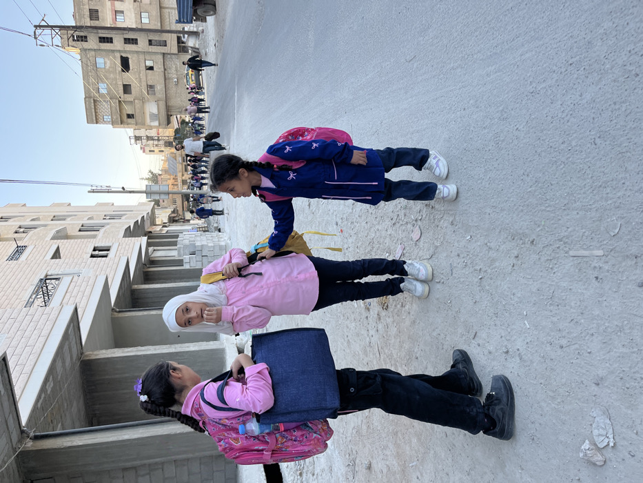 This image depicts a street scene in Mu'adammiyat ash-Sham, Syria, during daytime with clear skies.
In the foreground, three young females, likely school-aged children, are standing on a dirt and gravel path adjacent to a paved road. The girl on the left, with dark hair in a long braid adorned with a purple and white flower clip, wears a pink long-sleeved shirt, dark cargo pants, and black athletic shoes. She carries a pink backpack containing a visible water bottle and a dark blue tote bag. She faces slightly towards the center. The central girl wears a white hijab, a pink long-sleeved button-up shirt, dark blue jeans, and white athletic shoes, carrying a yellow backpack. Her right hand is near her mouth, and she looks directly towards the viewer. The girl on the right, the youngest, has dark hair in two braids and wears a dark blue long-sleeved tunic-style uniform with pink bow embroidery and trim, dark blue pants, and white and pink athletic shoes. She carries a pink backpack and her left hand is touching the central girl's yellow backpack, while she looks towards the central girl.
Behind the girls, partially constructed or renovated multi-story buildings with light-colored brick facades and exposed concrete are visible on the left. Stacked cinder blocks are present near the building base. On the right, older, weathered multi-story residential buildings line the street.
The paved road extends into the background, where a group of approximately 15-20 individuals, many appearing to be schoolchildren with backpacks, are walking away from the camera. Several adult males are also interspersed within the group. Utility poles with overhead wires and a street sign are visible along the road. A blue truck is partially visible on the far right. The overall light suggests morning or late afternoon.