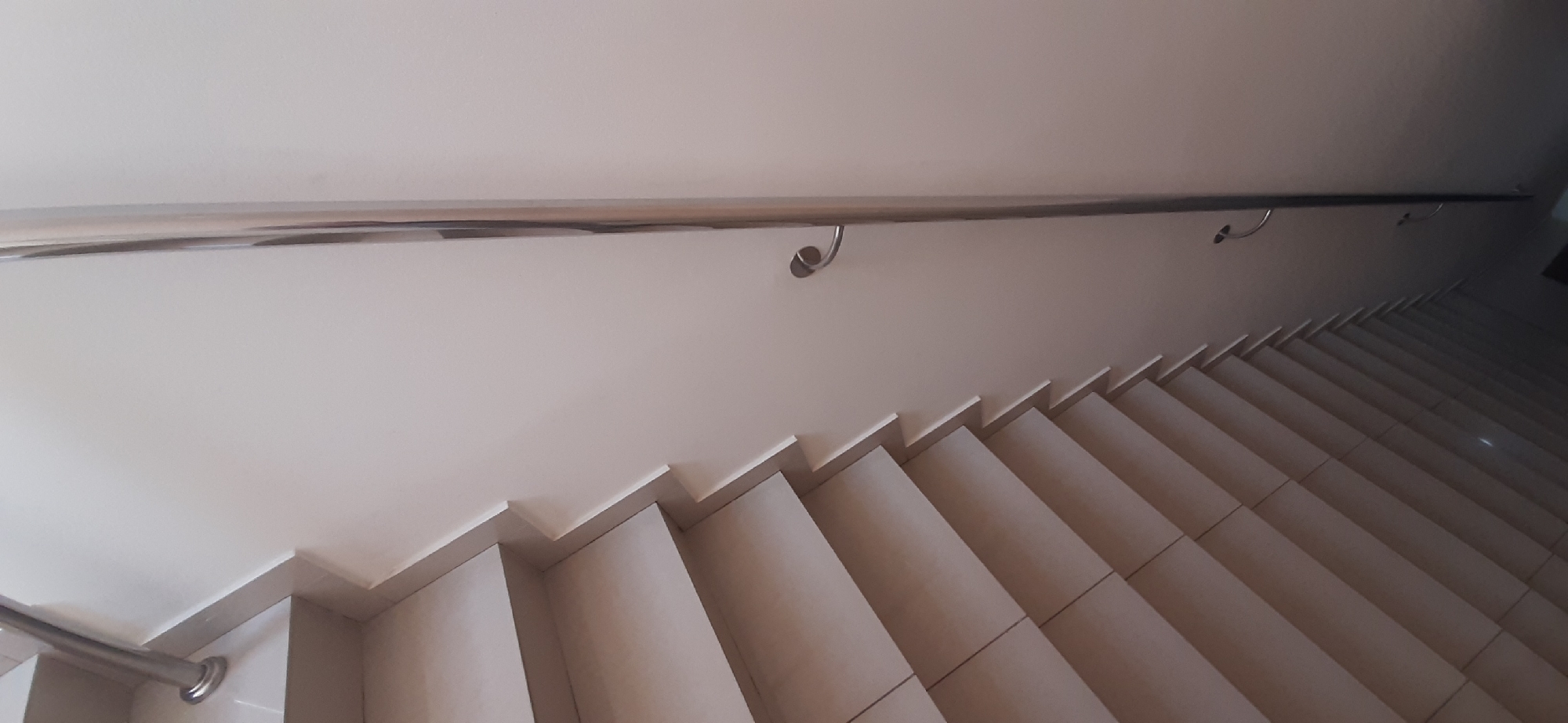 The image shows a view of a modern staircase with a polished metal handrail. The stairs are made of light-colored tiles with a matte finish, and they descend into shadow. The handrail is mounted on a plain, light-colored wall. There are three visible supports for the handrail, each with a curved design. The lighting suggests it is daytime, and the interior setting is likely a public building or a modern home. No people are visible in the image. The scene is static, with no activity taking place beyond the inherent functionality of a staircase. The overall impression is one of clean lines and minimalist design. The location context of Yessentuki, Russia, does not provide specific visual cues in the image itself, but suggests the architectural style might be found in that region. There is no visible text in the image.