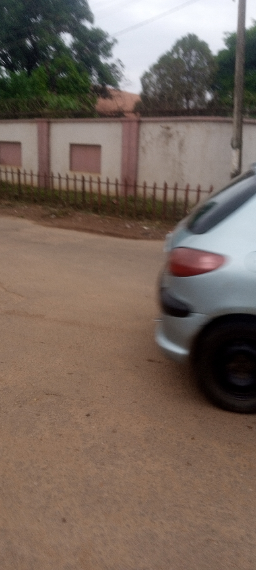The image depicts a blurred scene in what appears to be the Narayi area of Nigeria. A light-colored car, likely silver or gray, is moving, as indicated by the motion blur. Only the rear portion of the vehicle is visible, including the taillight, bumper, and rear tire. The road surface is a muted brown color, showing some signs of wear. In the background, a perimeter wall is seen, constructed of what appears to be concrete. The wall has a lighter central section and darker bordering vertical supports, with window-like impressions. Above the wall, greenery and trees are visible, partially obscuring the sky, which is overcast. A brown fence with vertical posts is between the street and the wall. A utility pole can be seen behind the car.