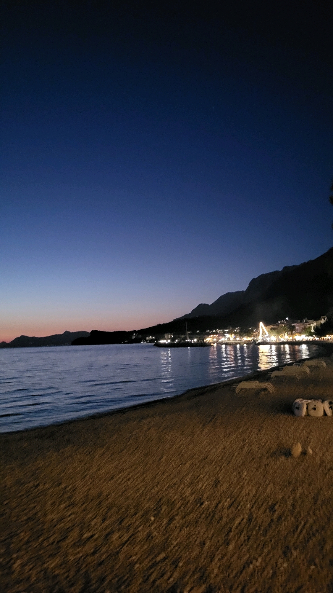 The image depicts a twilight scene on a beach in Općina Tučepi, Croatia. The sky transitions from a deep blue overhead to a gradient of purple and orange near the horizon, suggesting sunset or sunrise. A calm body of water stretches across the middle of the frame, reflecting the lights of a coastal town nestled along the base of a dark, mountainous silhouette. The town's lights are illuminated, creating a warm glow on the water.
The foreground is a sandy beach, with visible footprints and some scattered small stones. On the right side of the beach, several empty lounge chairs are arranged, some appearing to be made of white plastic. Near these chairs, a cluster of objects, possibly folded towels or cushions, are piled together. A palm tree is partially visible on the far right edge of the image. The overall atmosphere is tranquil and serene, capturing the end of the day in a coastal European setting.