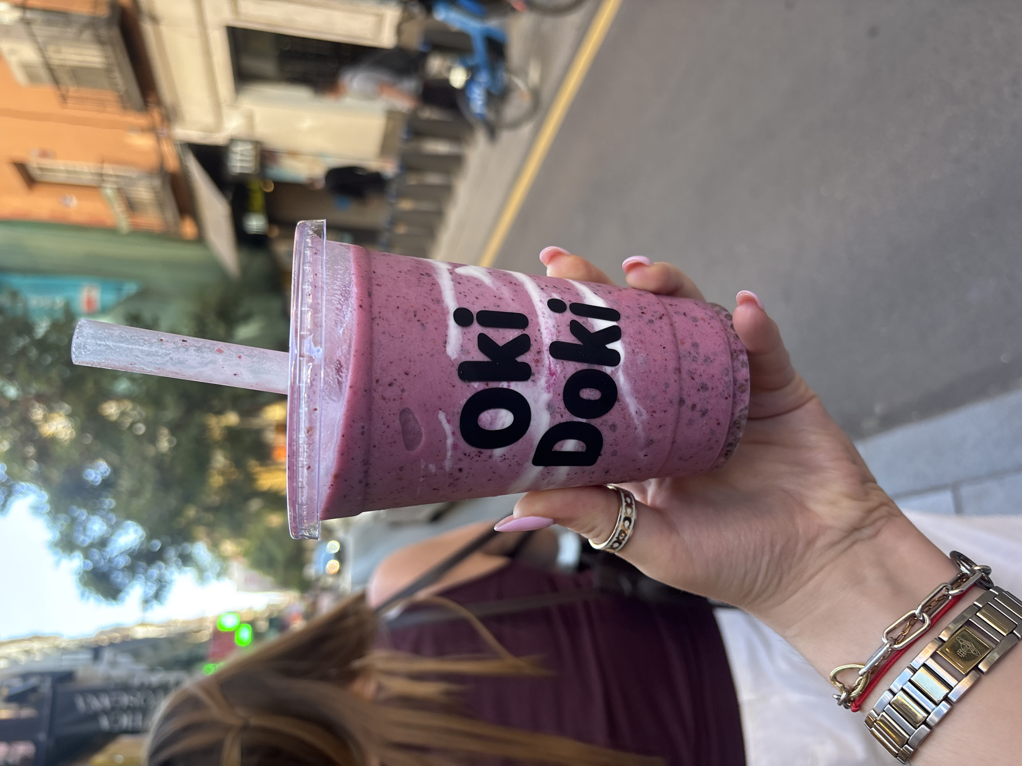 A person is holding a cup of a smoothie beverage outdoors. The drink has a purple-ish pink color with some white swirls, and a black "Oki Doki" logo printed on the side. A translucent straw is inserted into the cup. The person's hand is visible, with pink manicured nails and a ring on their finger. They are wearing a watch and other bracelets on their wrist. The background shows a street scene, with buildings, trees, and a bicycle visible. The city is Madrid, Spain.