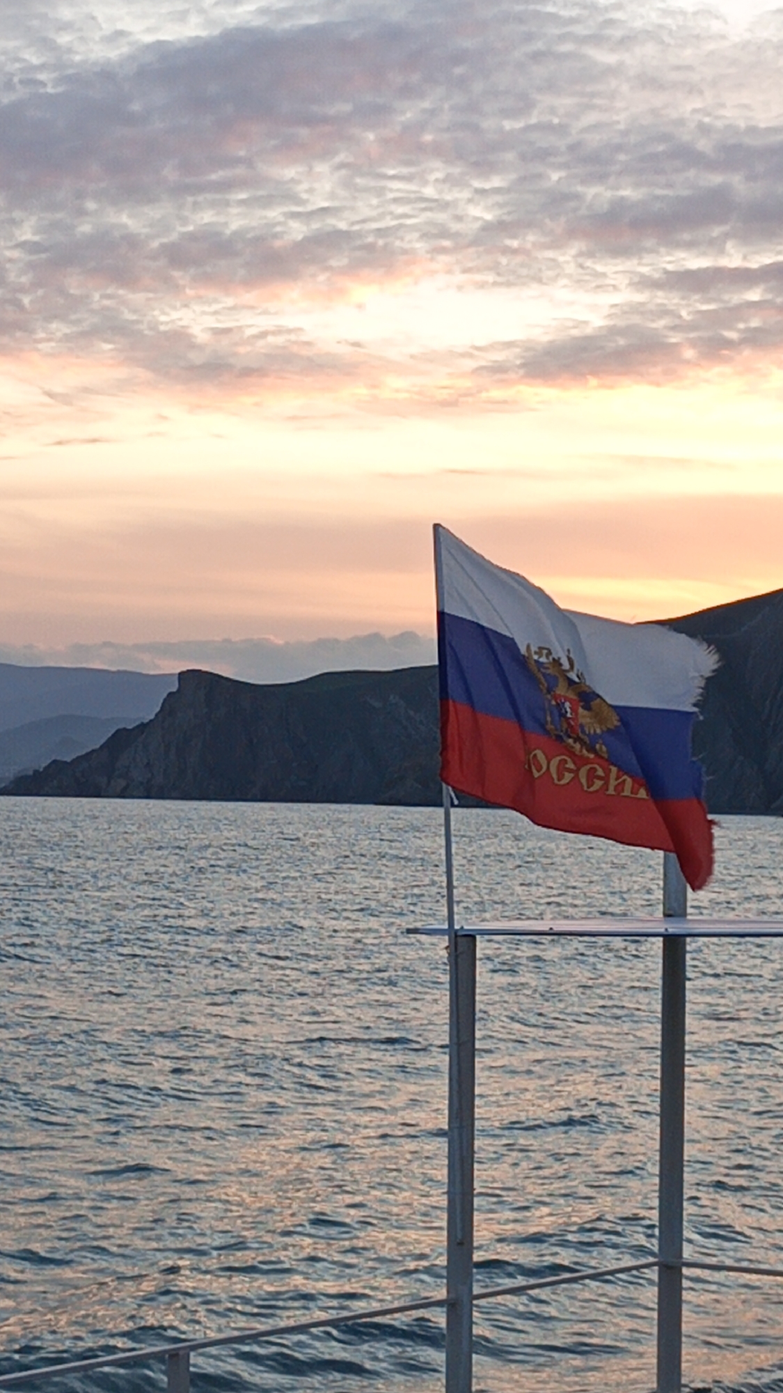 The image shows a partially tattered Russian flag waving on a metal pole in front of a body of water, likely the sea, with landmasses in the background. The sky is filled with clouds and a pinkish-orange hue, indicating sunrise or sunset. The flagpole appears to be part of a structure, possibly a pier or a boat. The flag displays the Russian tricolor (white, blue, and red) with the country's coat of arms and inscription "РОССИЯ." The scene is situated in Unknown, Unknown.