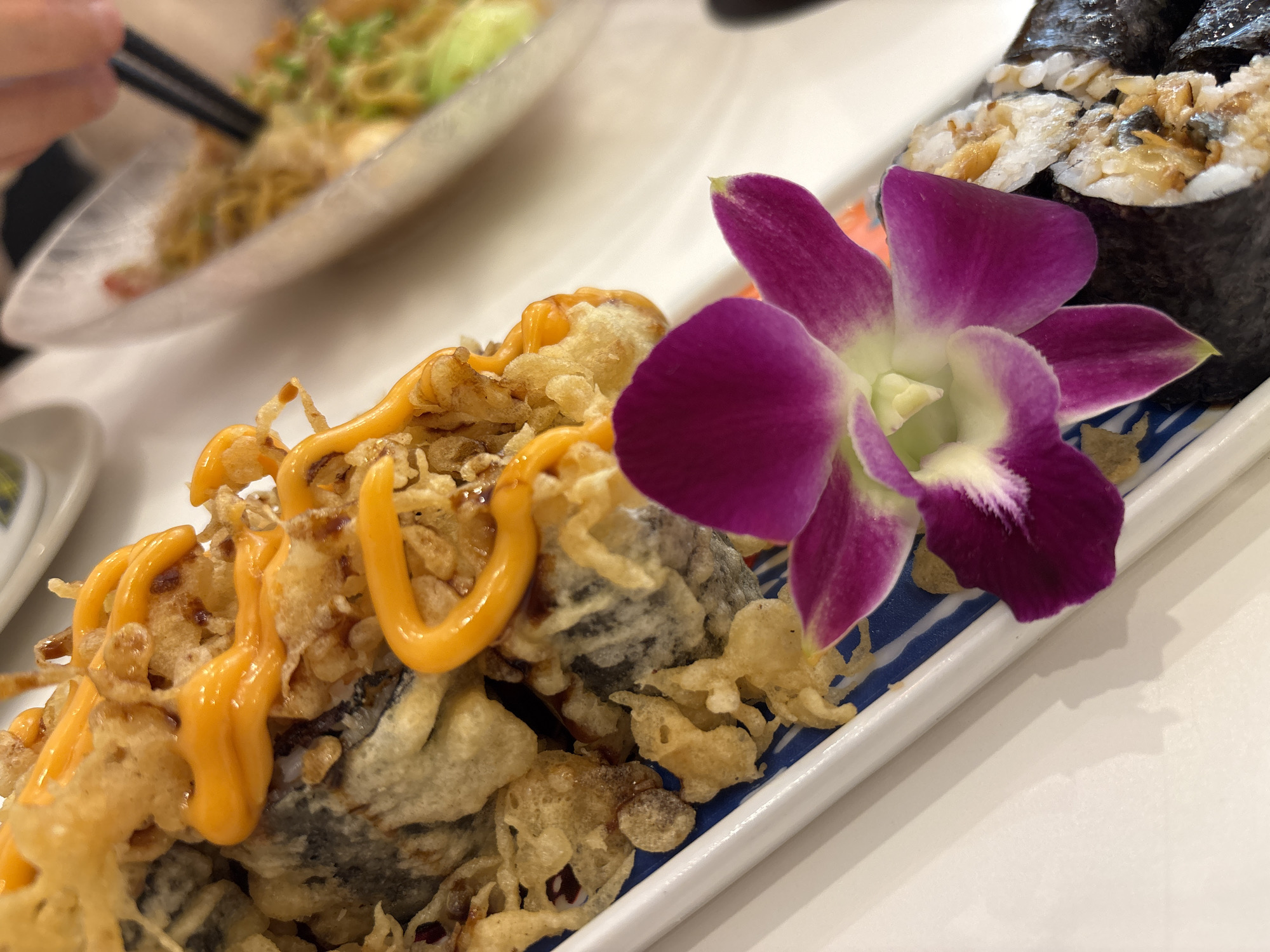 The image shows a close-up of a plate of food, likely taken at a restaurant. The dish features what appears to be tempura-battered food drizzled with an orange sauce, and next to it a roll of sushi, garnished with a purple orchid. In the background, a person can be seen using chopsticks to eat noodles from another dish. The scene is set on a white table, suggesting an indoor dining environment. The composition and plating of the food suggest a restaurant setting in Đà Nẵng, Vietnam.