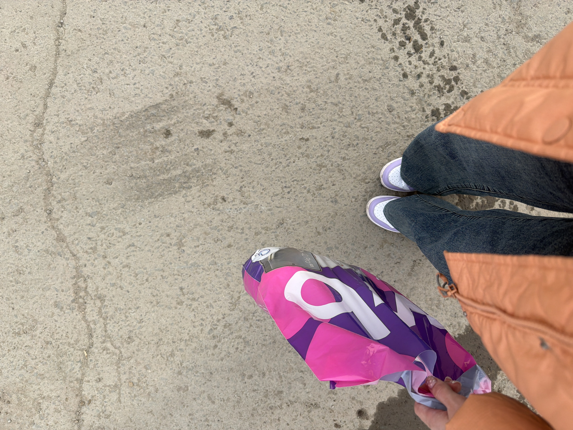 A ground-level perspective shows a person standing on a cracked concrete surface. The individual is wearing dark denim jeans, white sneakers with purple trim, and an orange puffy jacket. The person holds a shopping bag with purple and pink geometric designs in their hand. The bag's design suggests that the item may have been purchased from a store with a logo that includes the number "10," such as the "Honor 10" brand. The image was taken outside in a location with dappled sunlight, possibly in Berezniki, Russia.