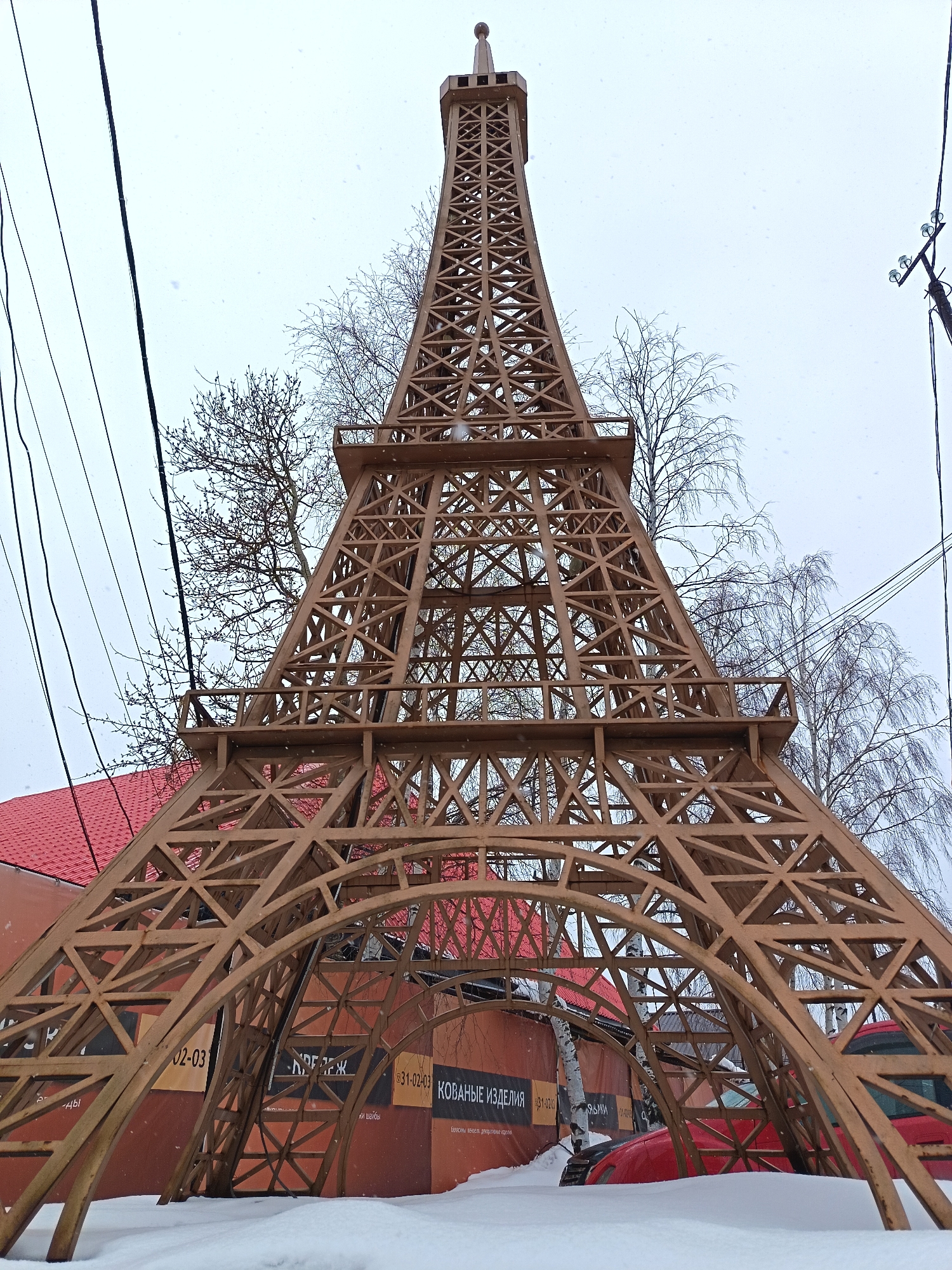 The image shows a scaled-down replica of the Eiffel Tower located in Nizhnevartovsk, Russia. It is a brown, metal structure standing in a snowy landscape, with a red-roofed building and bare trees visible in the background. Power lines are visible in the sky. Signs in Russian can be seen at the base of the Eiffel Tower. The ground is covered in snow.