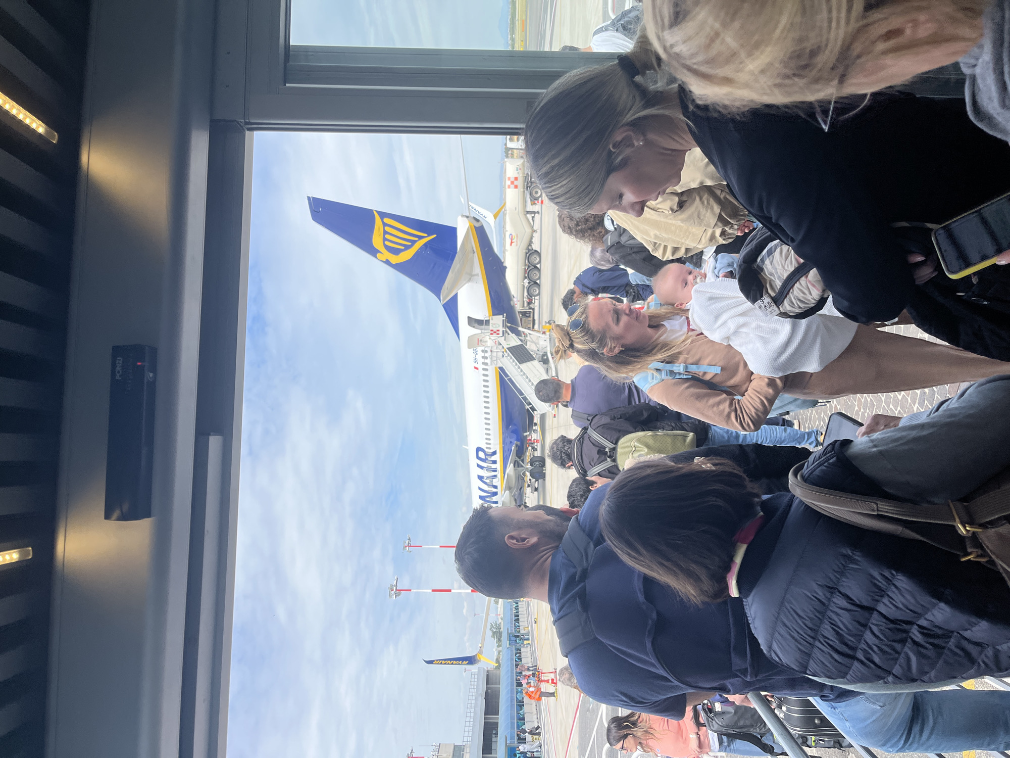 The image shows a crowded airport terminal gate in Rome, Italy. People are lined up, waiting to board a Ryanair airplane visible through the gate windows. The plane's distinctive blue and yellow tail is prominently displayed. Passengers are carrying luggage and personal items. A woman is holding a baby.