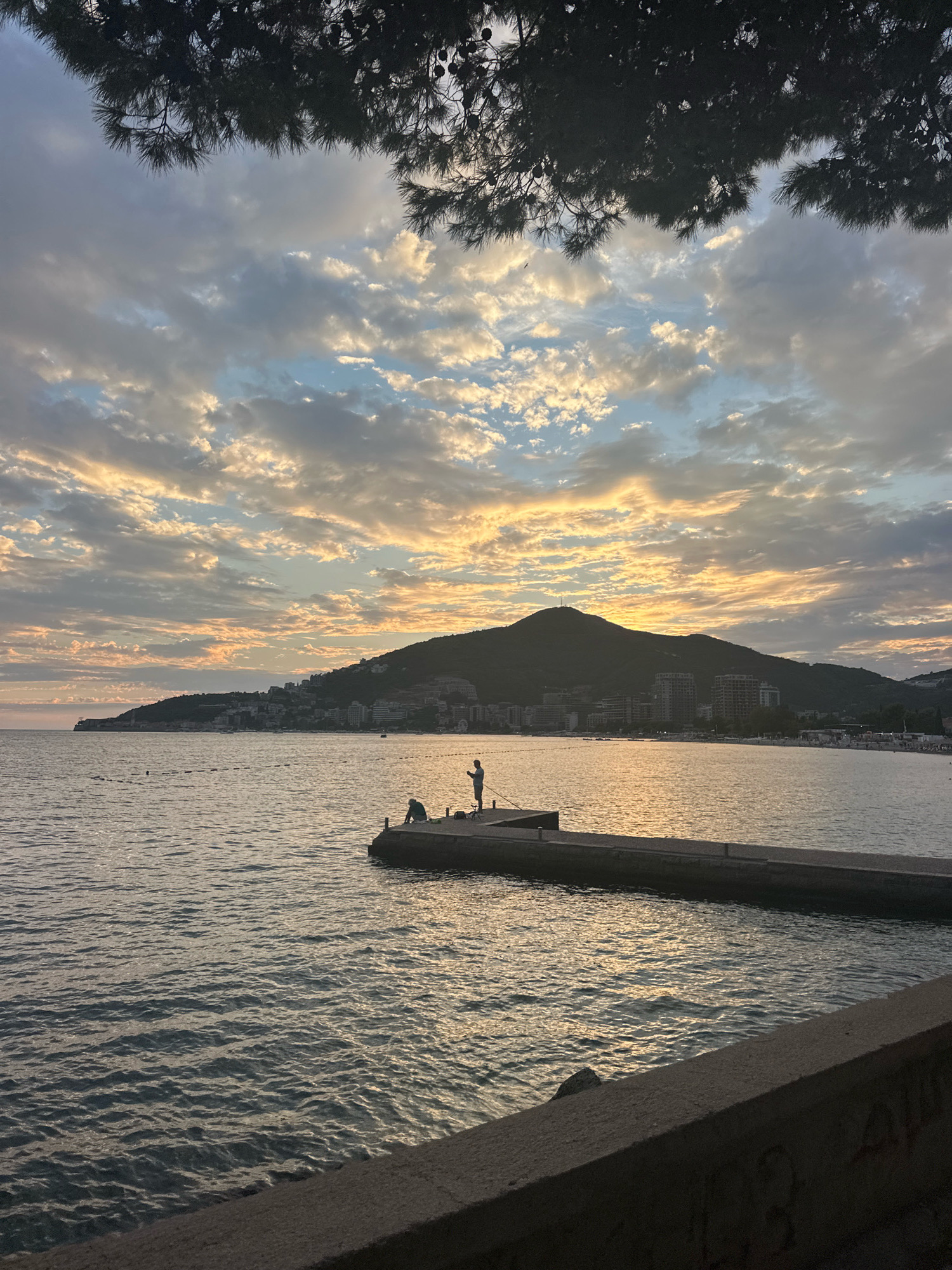 The image shows a sunset over the sea in Budva, Montenegro. In the foreground, a concrete pier extends into the water. Two figures are visible on the pier; one is sitting with their legs outstretched, while the other stands with a fishing rod. The sea is calm, with gentle waves reflecting the warm colors of the sky. In the background, a silhouette of a hill covered in buildings and vegetation rises from the coastline. The sky is filled with dramatic clouds, illuminated by the setting sun, casting golden and orange hues across the scene. Pine branches frame the top of the image, adding a natural element to the coastal view.