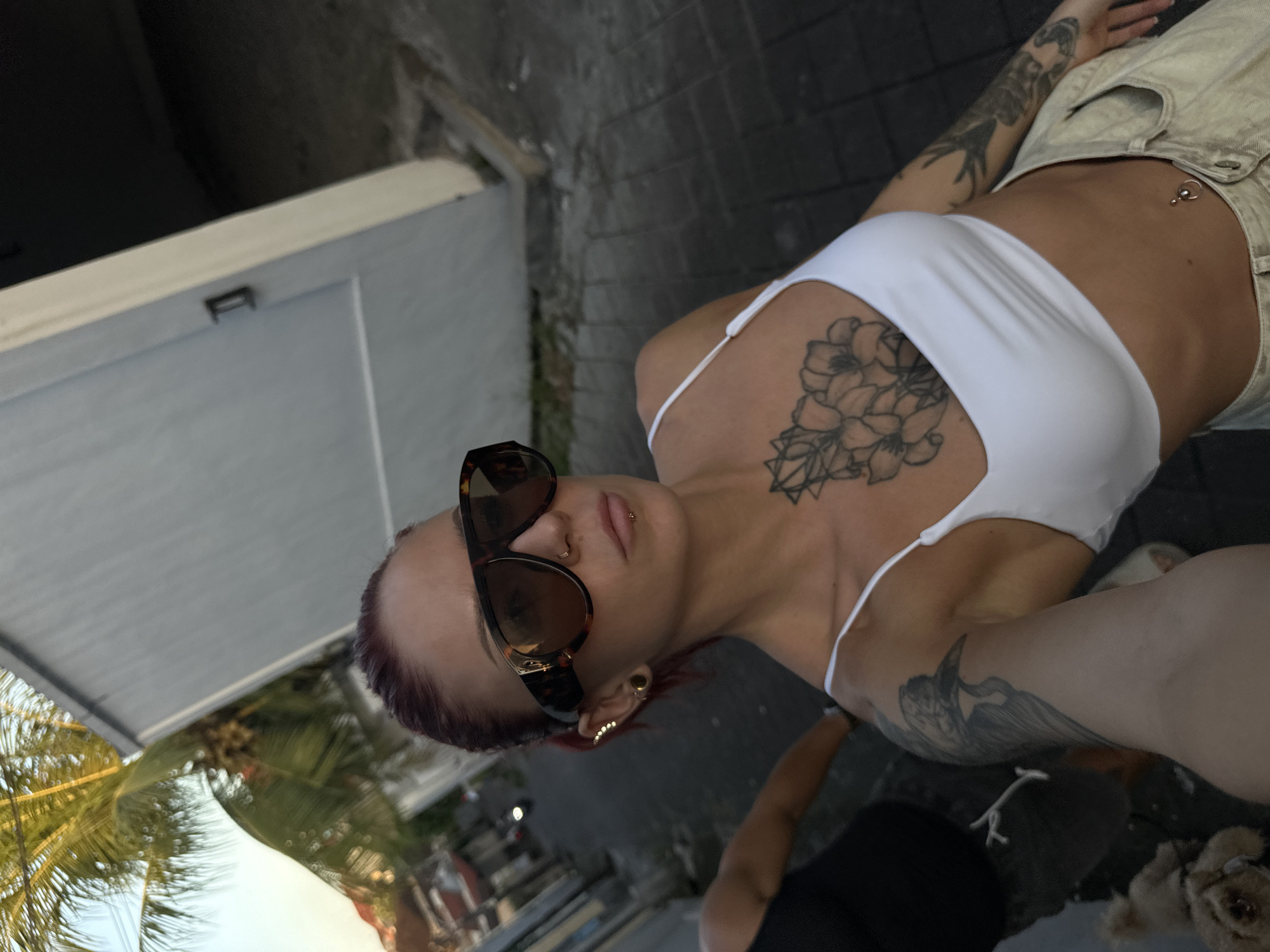 A woman with tattoos is posing outdoors. She has auburn hair, fair skin, and is wearing sunglasses, a white top, and beige shorts. She has body piercings on her nose, ear, and navel. Her tattoos include floral designs on her chest and arm, and other motifs on her arm.
The setting appears to be a street or walkway in an urban area. The background includes a building, trees (including palm trees), and other people in the distance. The presence of palm trees and the type of paving stones suggest a tropical location. Since North Kuta, Indonesia was mentioned as the location, this area is likely the location in the photograph.