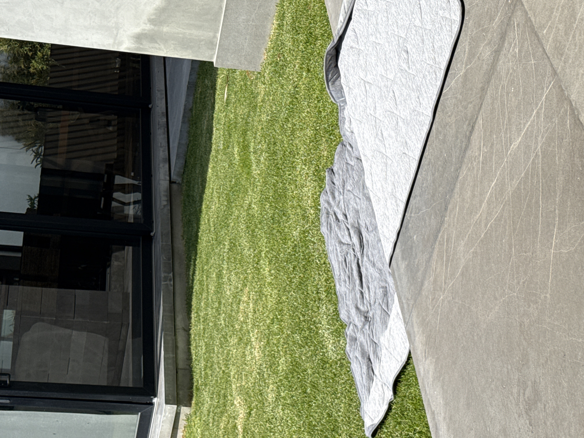 The image shows a patch of green grass next to a building with large glass windows. A gray tarp or cover is partially spread out on the grass and extends onto a section of concrete. The photograph was taken in San Bernardino Tlaxcalancingo, Mexico.