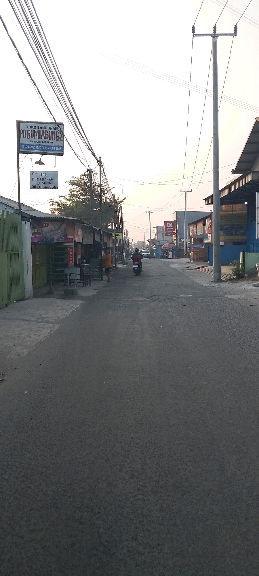 The image depicts a street scene in Satriajaya, Indonesia, with a view looking down a paved road lined with various businesses and buildings. The sky is bright, suggesting either early morning or late afternoon light.
On the left side of the road, there are several small storefronts with tin roofs and open fronts, displaying various goods and signs. One prominent sign reads "TOKO BANGUNAN PO BUMI AGUNG 2" with contact information below. Another sign, also on the left, indicates "ADA KONTRAKAN KOSONG" with a phone number. A green fence is visible to the far left.
The road itself is asphalt and appears to be in moderate condition, with some cracks and patches. In the middle distance, a motorcycle with a rider is traveling away from the camera. Further down the road, other vehicles and pedestrians are sparsely visible, suggesting a relatively quiet street.
On the right side of the road, there are larger buildings, some with modern architecture and awnings. Signs for businesses like "O! save mart" are visible. Utility poles with numerous electrical wires run along both sides of the street, dominating the upper portion of the image. Trees and vegetation can be seen in the background, interspersed with the structures. The overall atmosphere is that of a commercial area in a developing urban or semi-urban environment.